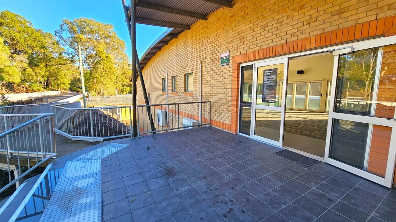 A brick building with a sloped roof on the right, featuring a glass door with a sign. The floor is tiled in dark grey. A metal railing lines the edge of the walkway, leading to a ramp on the left. In the background, there are tall trees and a clear blue sky. A small puddle and a single leaf are visible on the ramp.