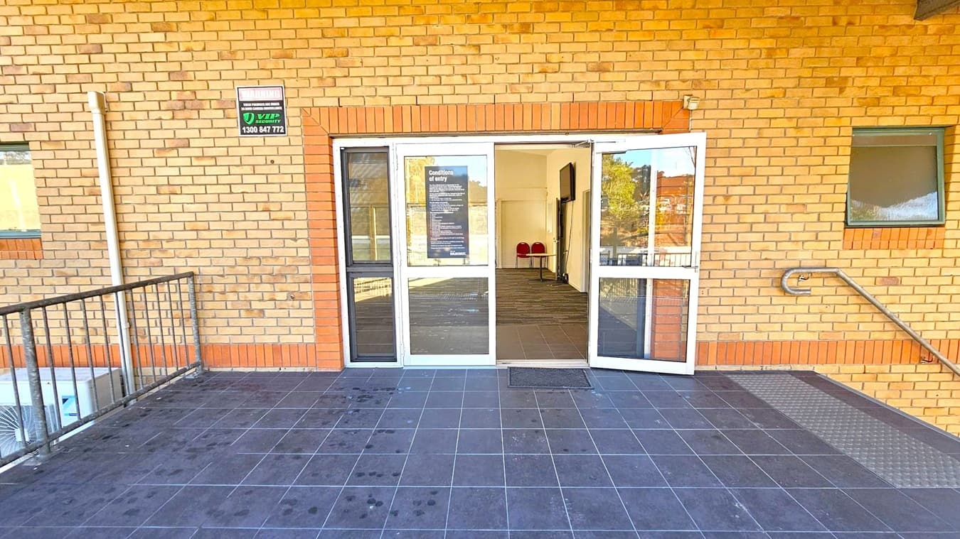 A brick building with a double glass door entrance in the centre, partially open. Above the door, a sign with security information is visible. Inside, a small table and two red chairs are seen. The floor outside is tiled in dark grey, with a metal ramp on the right. A metal railing is on the left, and a small window is on the right side of the wall.