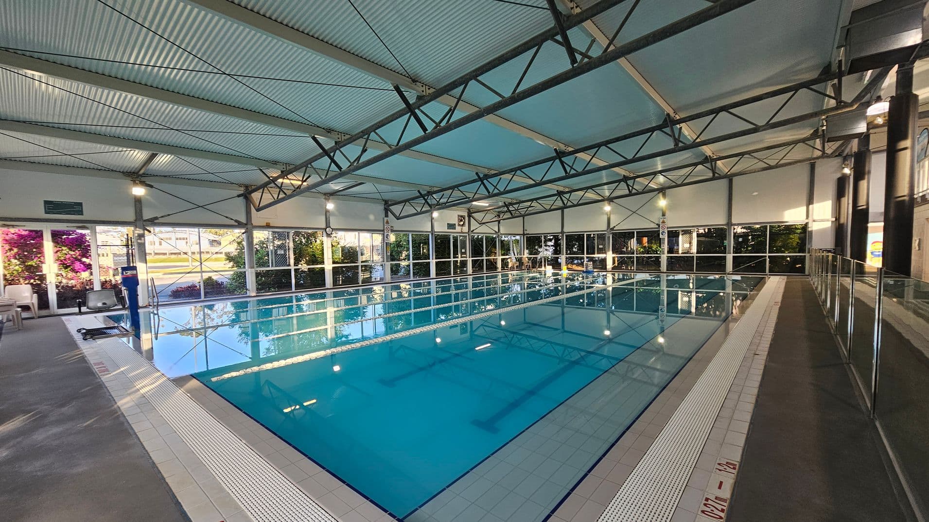 On the left, a concrete walkway with beige seating near large glass windows and vibrant purple flowers. In the middle, a rectangular swimming pool with bright blue water is divided by lane markings, surrounded by white tiled edges. On the right, a glass balustrade runs alongside a grey non-slip floor. The ceiling is corrugated metal with exposed triangular beams and fitted with suspended lights.