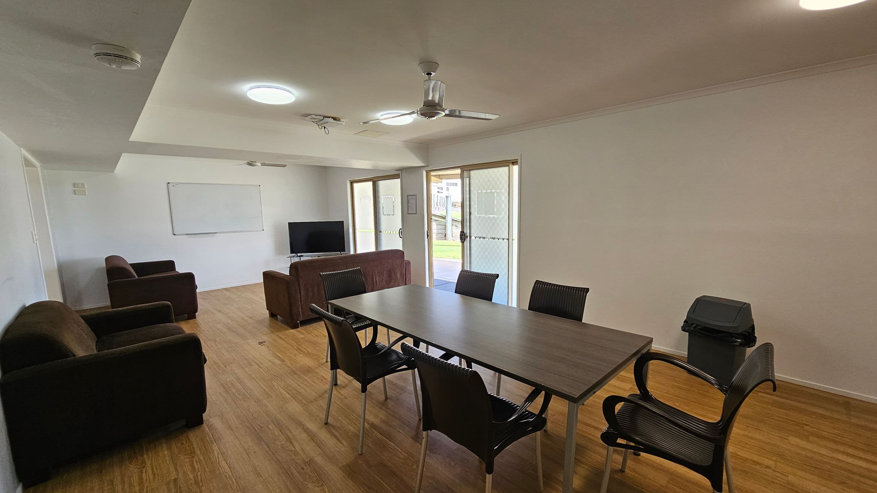 A room with wooden flooring and a white ceiling featuring recessed lighting and a ceiling fan. On the left, there are two brown armchairs and a brown sofa facing a television on a stand. A whiteboard is mounted on the wall behind them. In the centre, a rectangular table is surrounded by six black chairs. On the right, there are glass doors leading outside, and a small bin is placed near the wall.