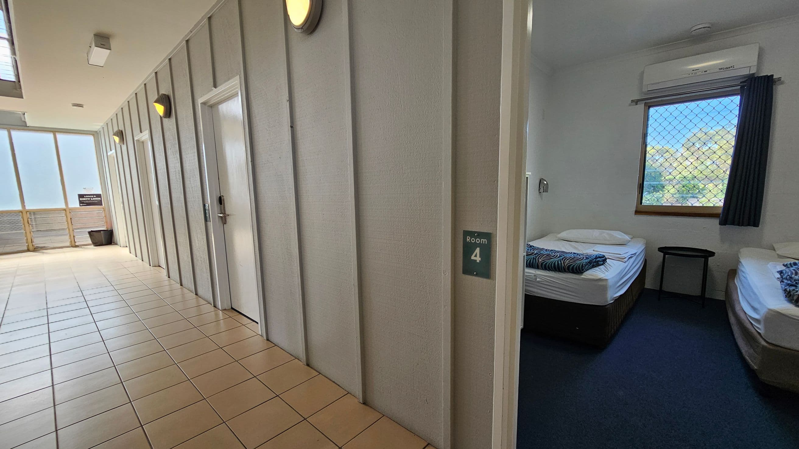 A hallway with light-coloured walls and tiled flooring. On the left, there are several closed doors with wall-mounted lights above them. On the right, an open door labelled "Room 4" reveals a room with two single beds, each with folded linens. The room has a window with a security screen and a curtain, and an air conditioning unit above. A small table is positioned between the beds.