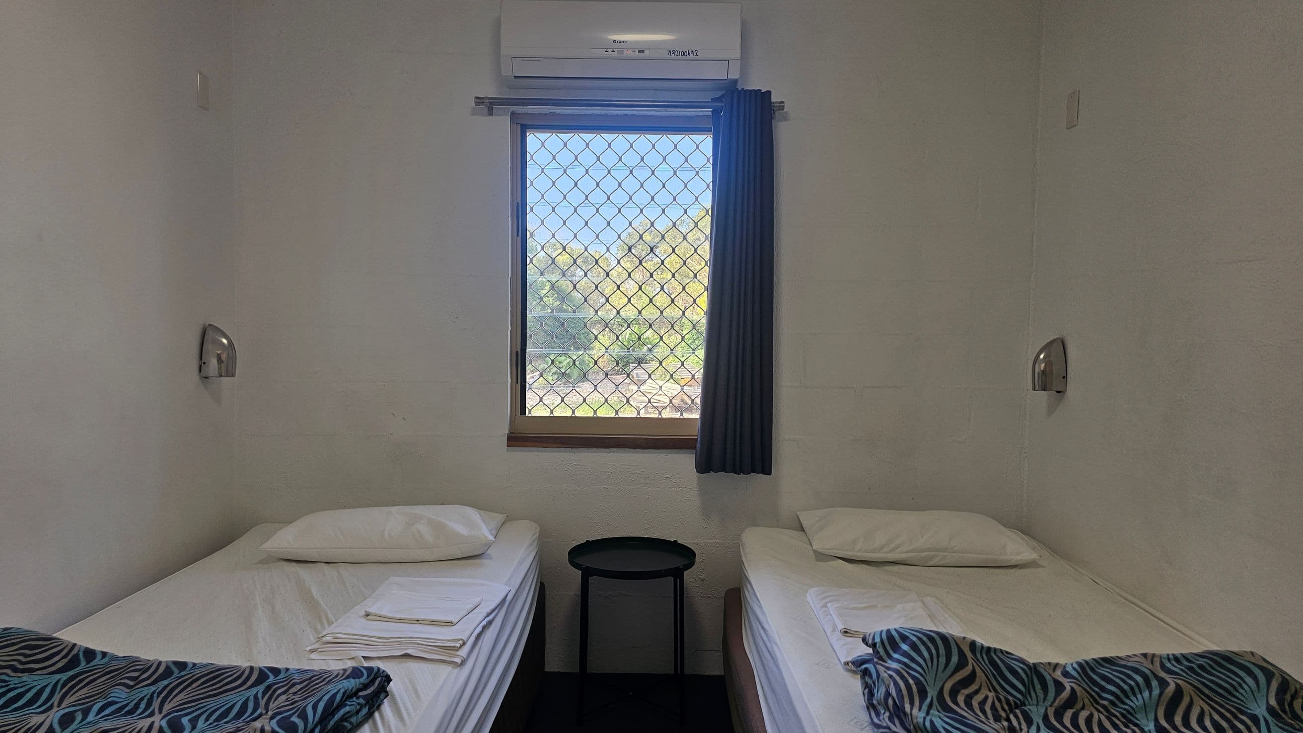A small room with two single beds on either side, each with a pillow and a folded blanket featuring a blue and black pattern. A small round black table is positioned between the beds. Above the table is a window with a mesh screen, partially covered by a dark curtain, showing greenery outside. An air conditioning unit is mounted above the window. The walls are plain, and there are small wall-mounted lights above each bed.