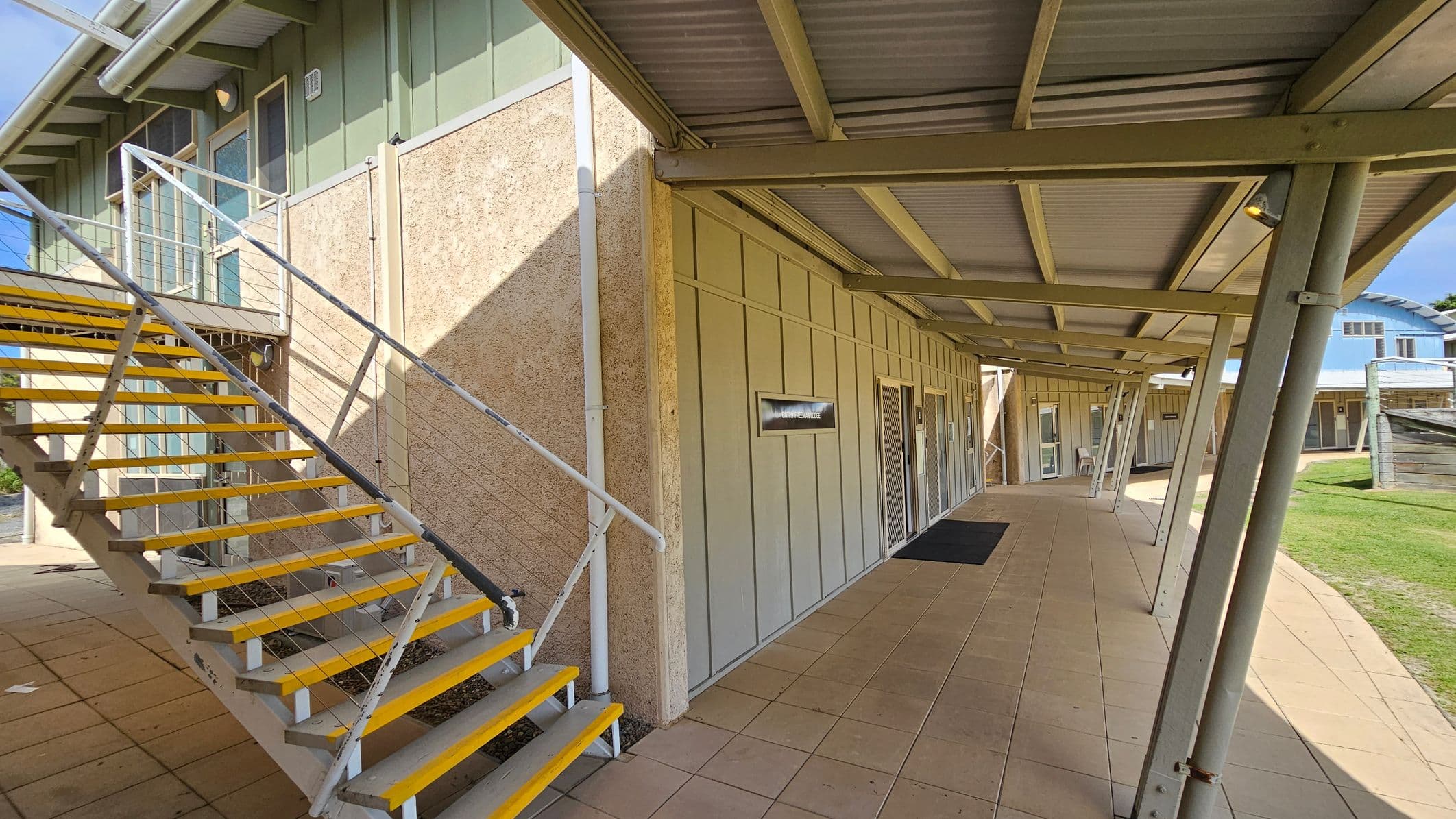 A covered walkway with a metal roof and supporting beams on the right. The floor is paved with light-coloured tiles. On the left, a metal staircase with yellow-edged steps leads to an upper level. The building walls are a mix of textured and panelled surfaces, with small windows and doors along the corridor. In the background, a grassy area and another building with a blue roof are visible.