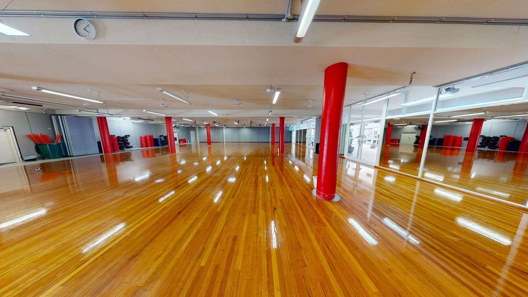 On the left, storage bins with red objects and stacked equipment are along a grey wall. In the middle, a spacious room with polished wooden flooring and evenly spaced red pillars extends to the background, where a grey folding partition wall is visible. On the right, a wall of floor-to-ceiling mirrors reflects the room, with glass doors leading to other spaces. The beige ceiling has suspended fluorescent lights, exposed piping, and ceiling fans.