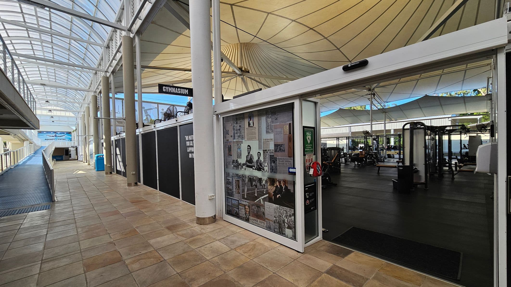 A corridor with a tiled floor and a glass ceiling on the left, leading to an area marked "Aquatic Suite." On the right, there's an entrance to a gymnasium with a glass door displaying various newspaper clippings and images. Inside the gym, exercise equipment is visible on a dark floor. The ceiling above the gym is covered with large, white, tent-like structures. A blue recycling bin is positioned along the corridor.