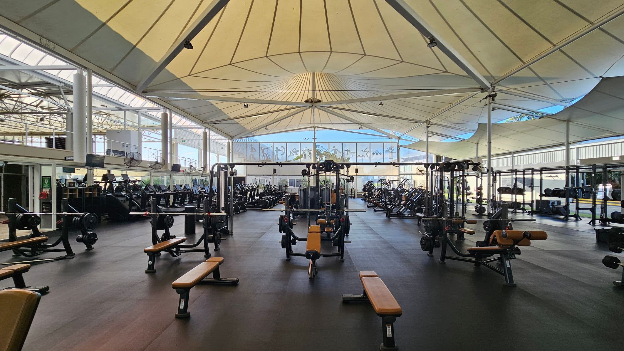 A spacious gym interior with a high, tent-like ceiling. In the foreground, there are several weight benches and weightlifting equipment. The middle area is filled with various exercise machines, including treadmills and ellipticals. The background features large windows with frosted designs, allowing natural light to filter in. The floor is dark and rubberised, suitable for gym activities. The ceiling has a geometric pattern with supporting beams.