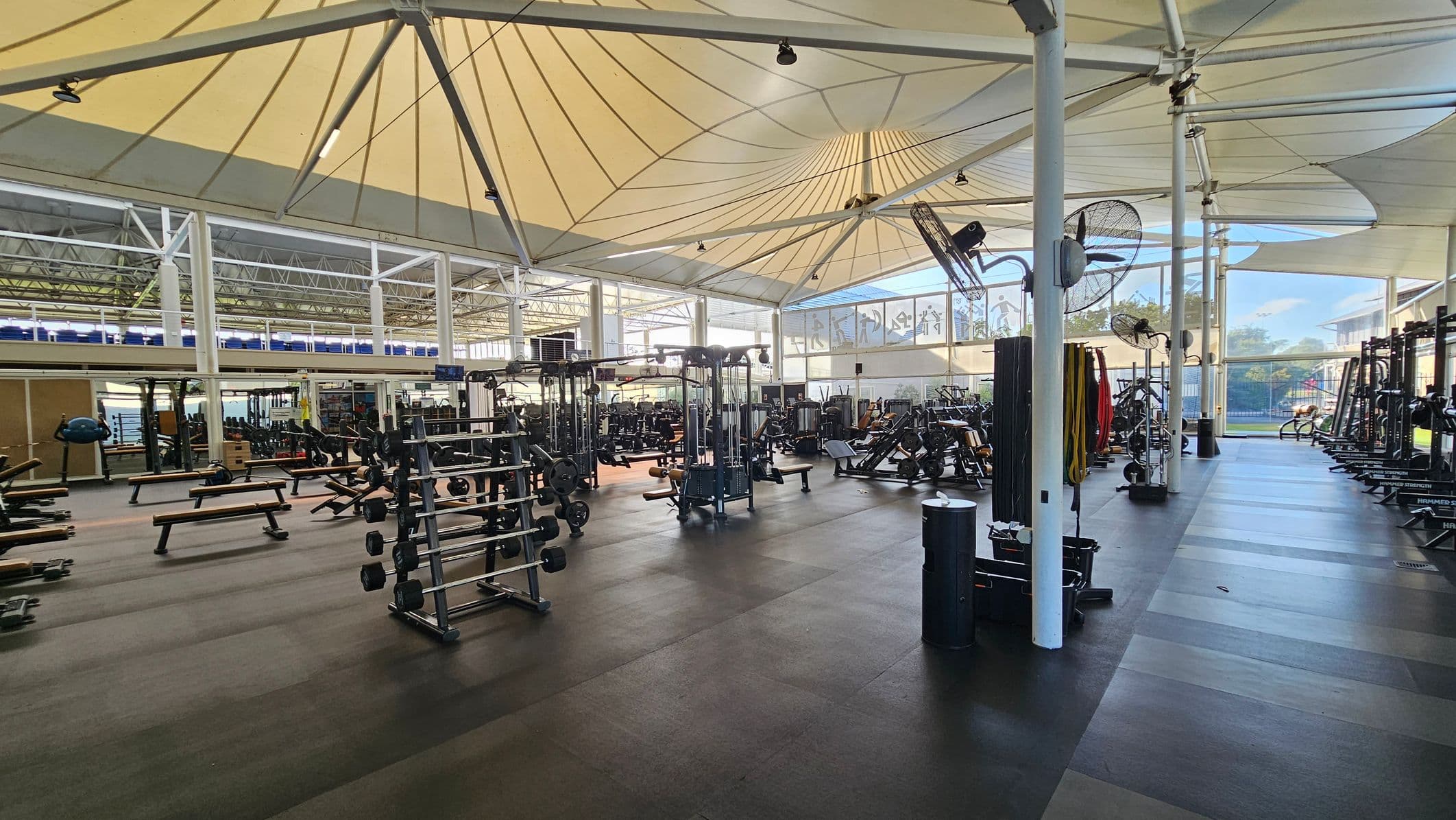 A spacious gym with a high, tent-like ceiling supported by white beams. On the left, there are weight benches and racks of dumbbells. In the centre, various weight machines and equipment are arranged. On the right, more exercise machines and racks are visible. The floor is covered with dark rubber mats. Large windows in the background allow natural light to fill the space, and several fans are mounted on the beams.