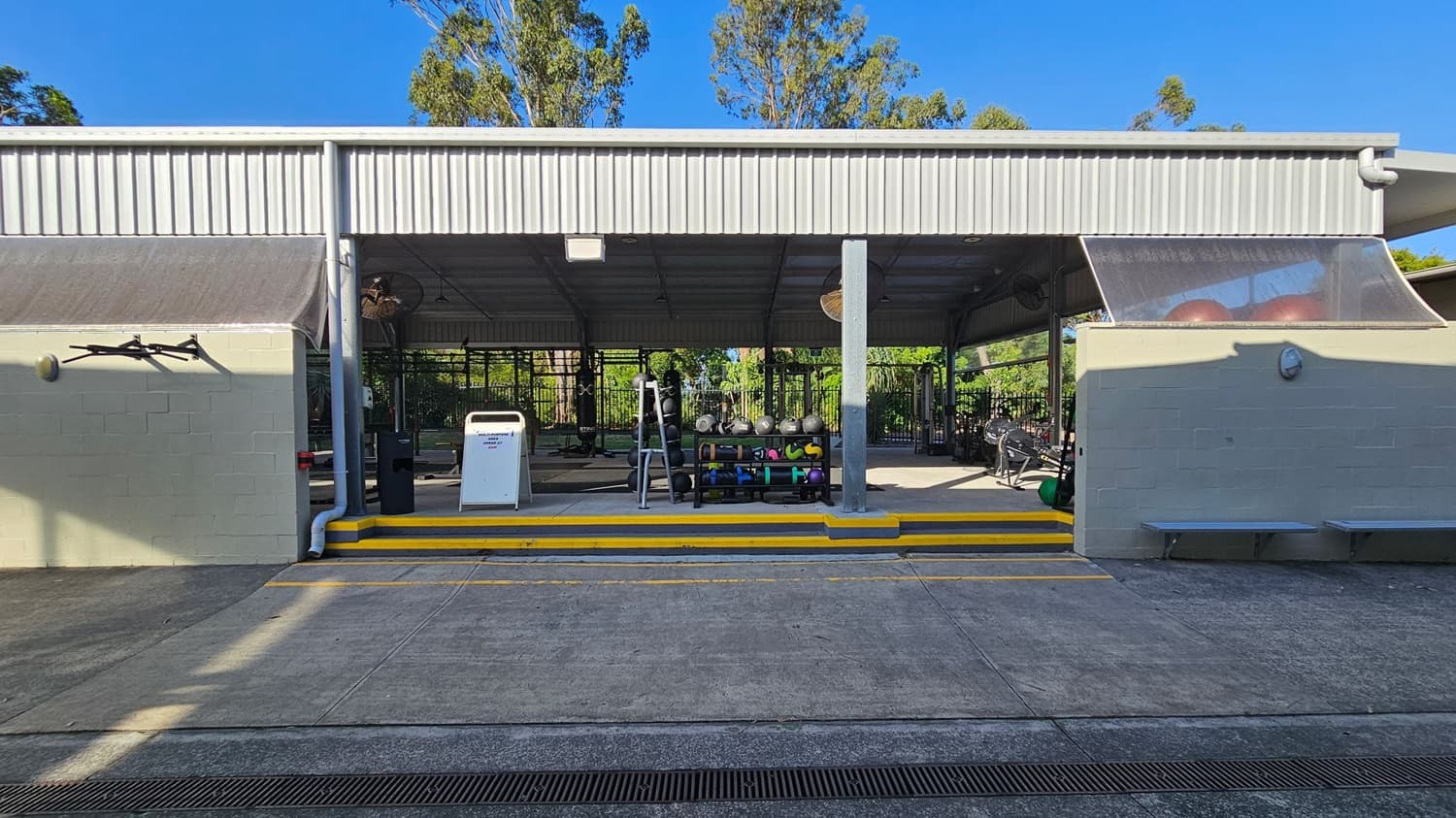 An outdoor gym area with a metal roof and open sides. In the centre, there are various gym equipment, including kettlebells and medicine balls on racks. A white signboard is placed near the entrance. The floor is concrete with yellow-striped steps leading up to the gym area. On the left, a wall-mounted light fixture is visible. The background features green trees and foliage.