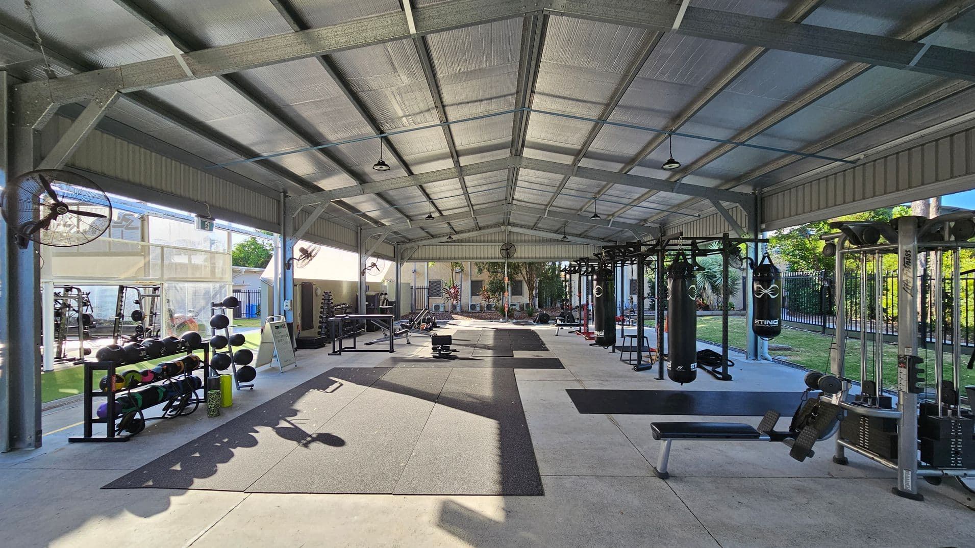 An open-air gym with a metal roof and concrete floor. On the left, there are racks of medicine balls and weights. In the centre, various exercise equipment is arranged, including benches and weightlifting stations. On the right, there are punching bags and cable machines. Large industrial fans are mounted on the walls, and the area is surrounded by greenery and a fence in the background.