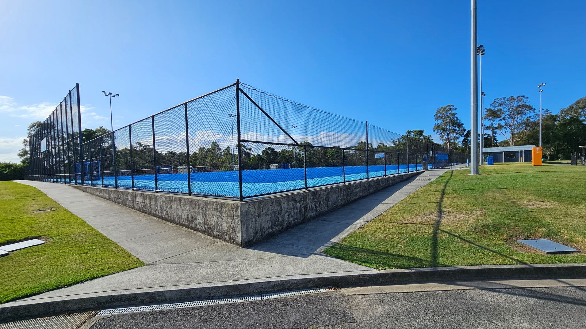 A blue sports court is enclosed by a tall chain-link fence, situated on a raised concrete platform. To the left, a sloped concrete path runs alongside a grassy area. On the right, a large metal floodlight pole stands on a grassy field, with a small building and bins in the distance. Trees are visible in the background, and the sky is clear.