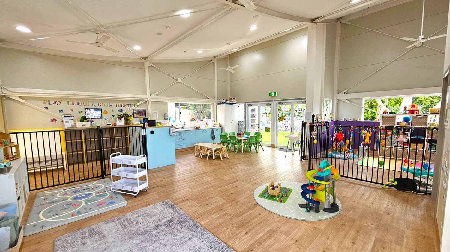 A spacious, well-lit playroom with wooden flooring. On the left, there is a play kitchen and a small white trolley on a grey rug. In the centre, a light blue cabinet with a countertop and a small table with green chairs. The right side features a fenced play area with colourful toys and a storage unit. Large windows and doors in the background open to a green outdoor area. The ceiling is high with visible beams and ceiling fans.