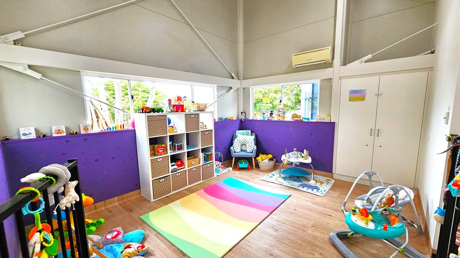 A children's playroom with a light wooden floor and a high ceiling. On the left, a black railing is adorned with colourful toys. In the centre, a white shelving unit holds various toys and books, with a rainbow-coloured mat in front. A blue chair with a cushion and a basket of toys is nearby. On the right, a baby activity centre and a bouncer are placed on a mat with animal designs.