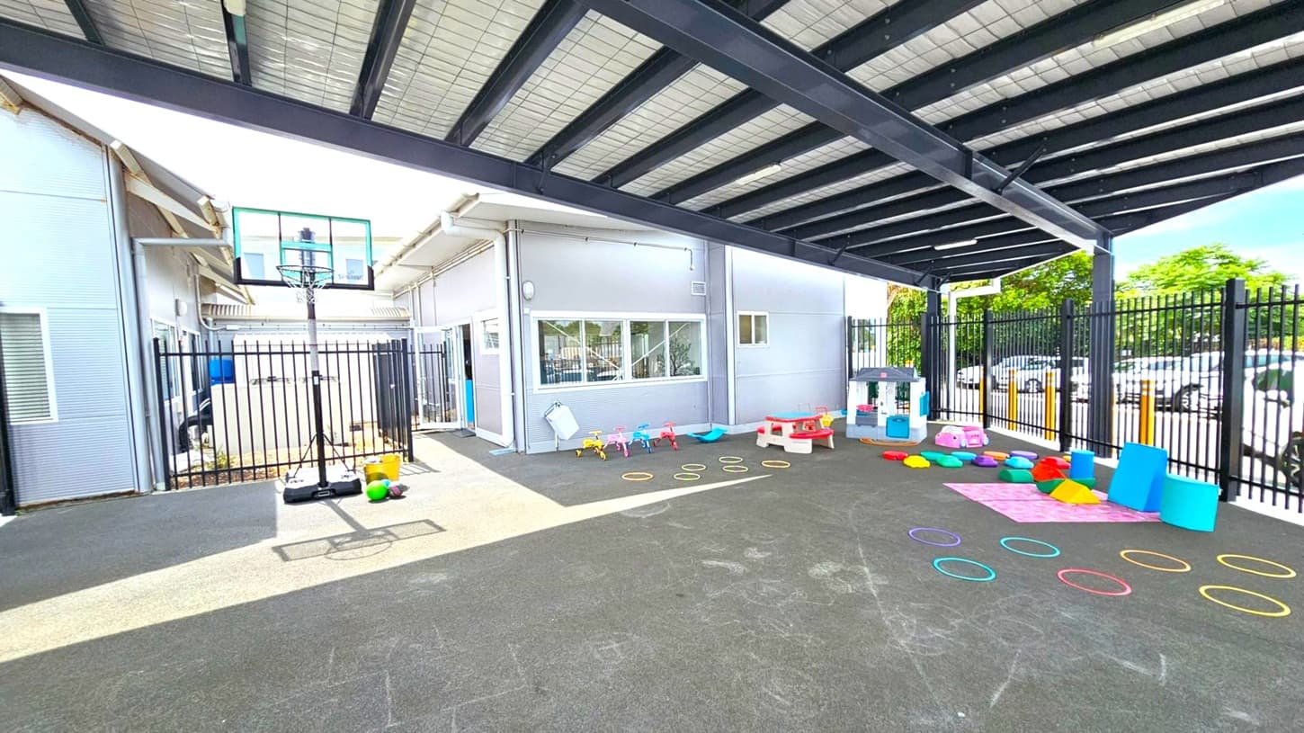 An outdoor play area with a black and grey rubber floor. On the left, there's a basketball hoop and some balls. In the centre, colourful hoops are arranged on the ground. On the right, a small playhouse, a red picnic table, and various colourful play structures are visible. The area is enclosed by a black metal fence, and a building with large windows is in the background. The ceiling is a metal structure with beams.