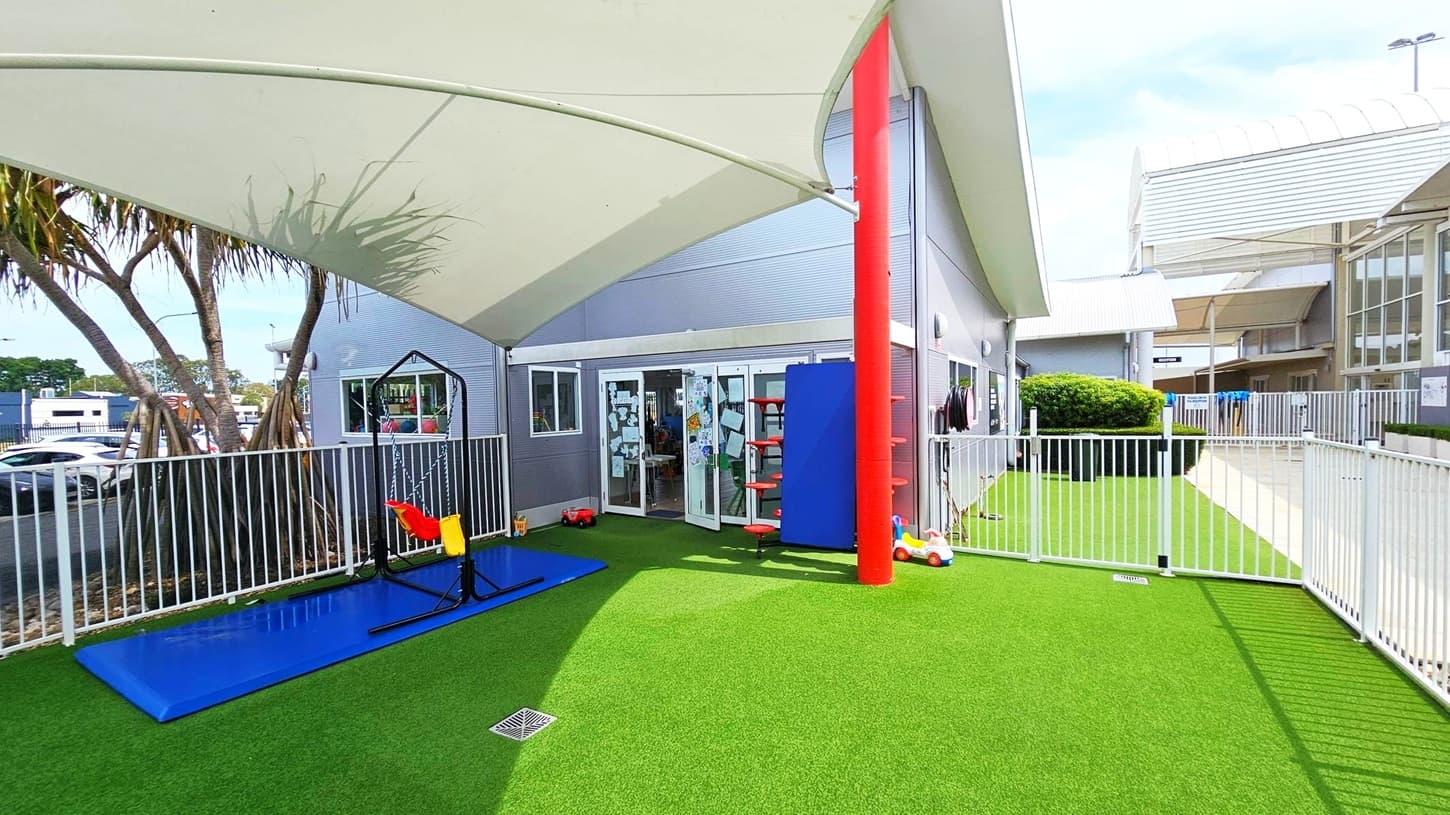 A playground area with artificial grass and a white fence. On the left, there's a blue mat with a swing set featuring red and yellow seats. A large shade sail covers the area, supported by a red pole. In the centre, glass doors lead into a building with children's artwork displayed. On the right, a small toy car is near the fence, and a bush is visible in the background. The ceiling is a white fabric canopy.
