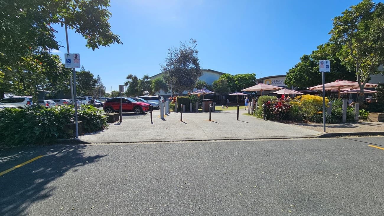 A car park with several vehicles, including a red car on the left. In the centre, there are bollards and a pathway leading to a building partially visible in the background. On the right, there are outdoor seating areas with red and white striped umbrellas, surrounded by greenery and colourful plants. Parking signs are visible on both sides. Trees provide shade throughout the area.