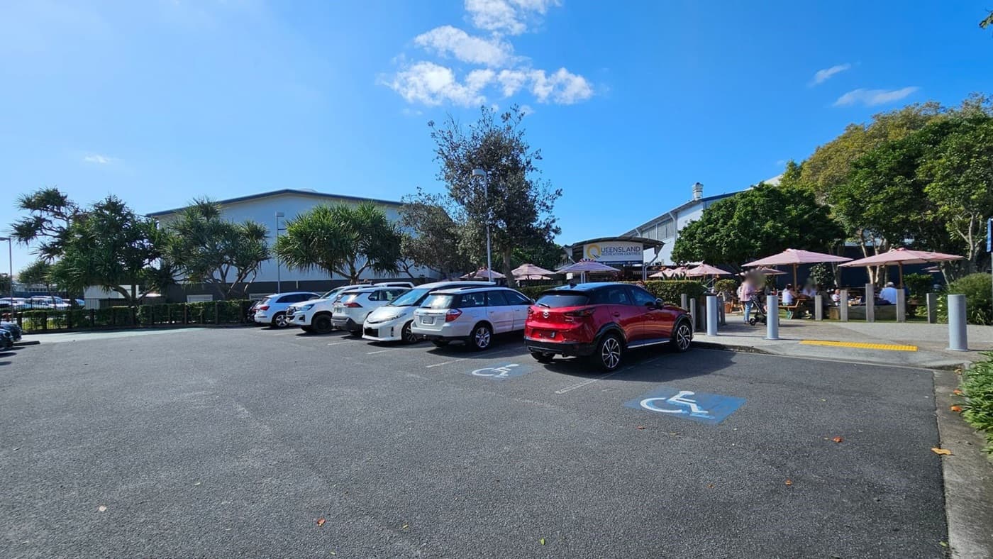 A car park with several vehicles, including a red car in a disabled parking space. On the right, there are outdoor tables with umbrellas and people seated, likely a café or restaurant area. A sign reads "Queensland" in the background. Trees and shrubs are scattered around, providing some greenery. The ground is asphalt, and there are bollards near the seating area.