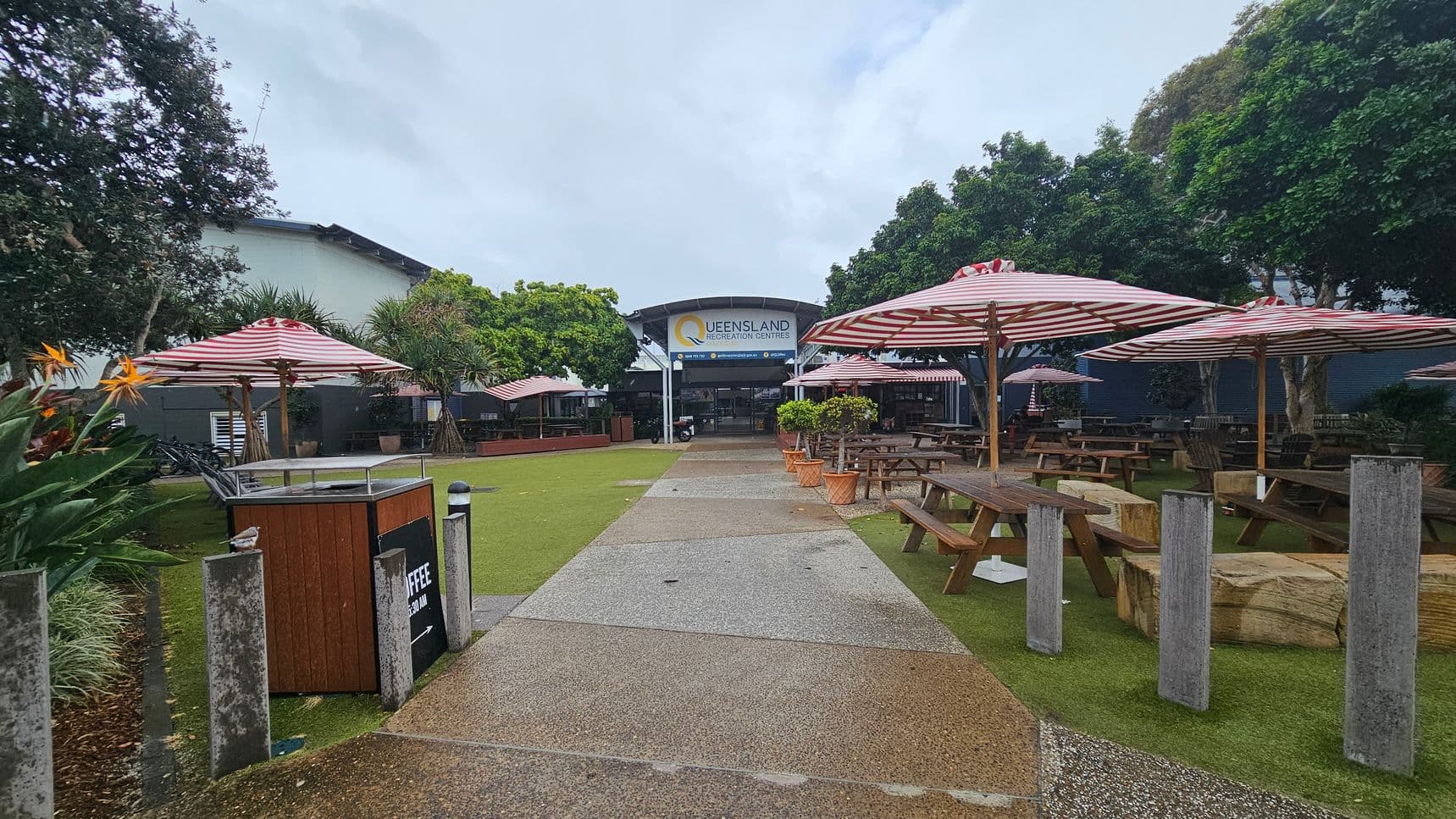A pathway leads to a building with a sign reading "Queensland Recreation Centres" in the background. On both sides of the path are picnic tables with red and white striped umbrellas. The area is surrounded by lush green trees and plants, including bird of paradise flowers on the left. The ground is a mix of grass and concrete, and there are several concrete bollards along the path.