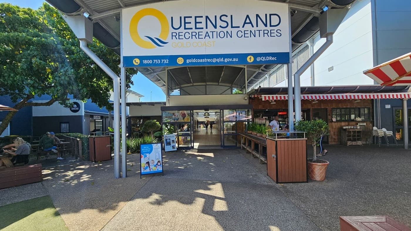 Entrance to Queensland Recreation Centres, Gold Coast. A large sign is overhead. On the left, people sit at wooden benches under a tree. On the right, a café with a red and white striped awning and a counter with plants. The centre has glass doors leading inside. The ground is paved, and the ceiling is metallic with beams.