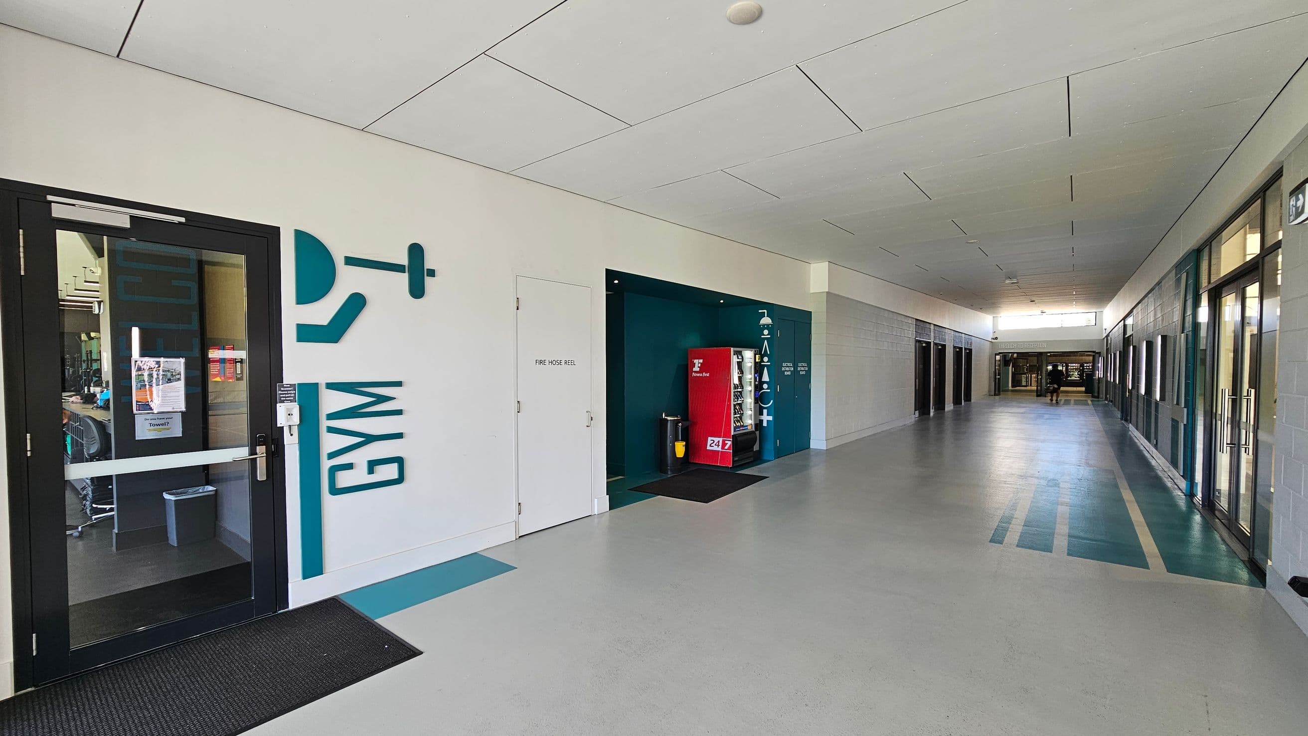 A corridor with a light grey floor and a white ceiling. On the left, there's a glass door with "GYM" written beside it, leading to a gym area. Next to it is a door labelled "FIRE HOSE REEL". Further along, there's a red vending machine in a recessed area. On the right, there are several glass doors and windows. In the background, two people are walking towards the end of the corridor, where more doors and windows are visible.