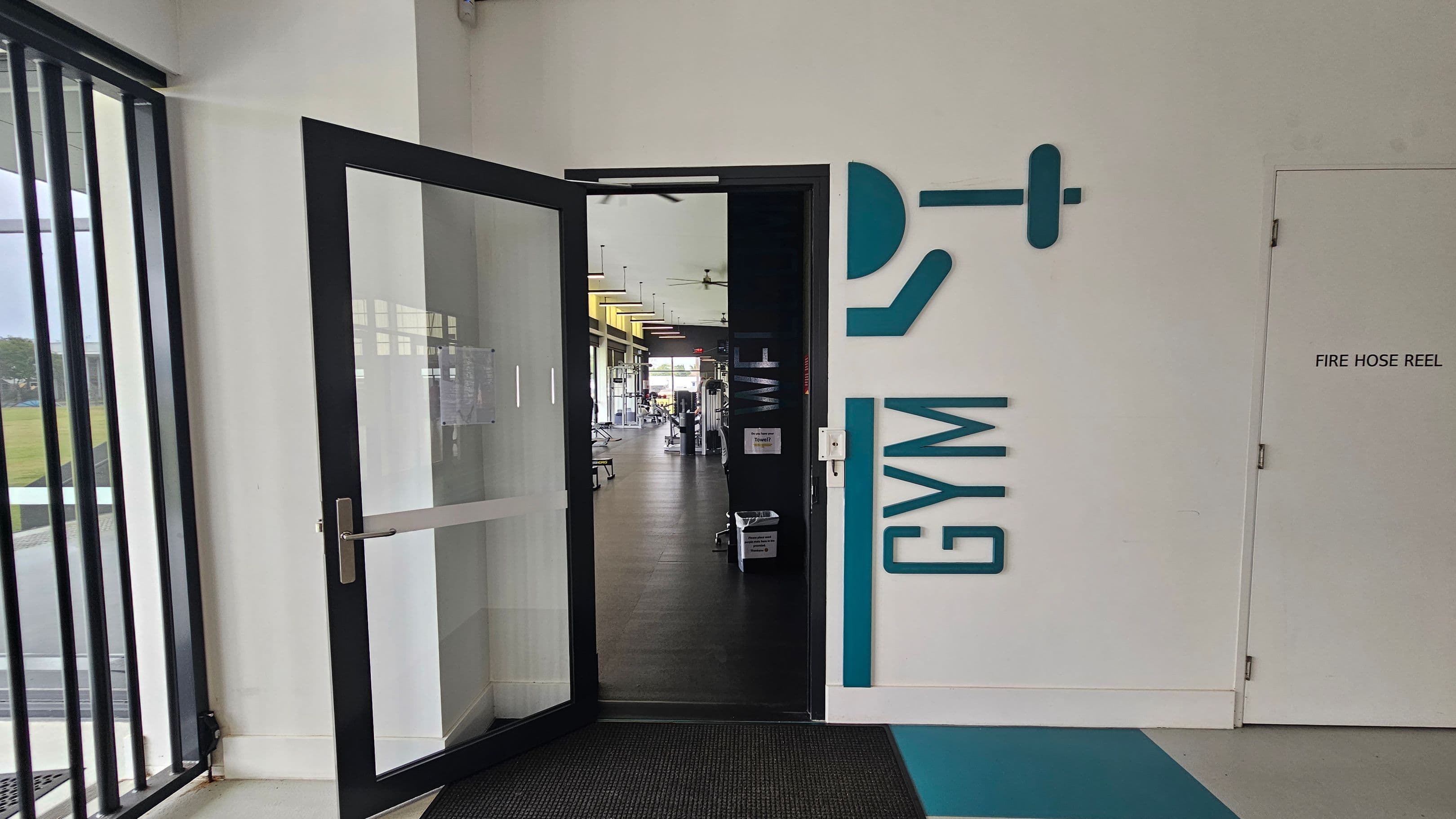 A corridor with a light grey floor and a white ceiling. On the left, there's a glass door with "GYM" written beside it, leading to a gym area. Next to it is a door labelled "FIRE HOSE REEL". Further along, there's a red vending machine in a recessed area. On the right, there are several glass doors and windows. In the background, two people are walking towards the end of the corridor, where more doors and windows are visible.