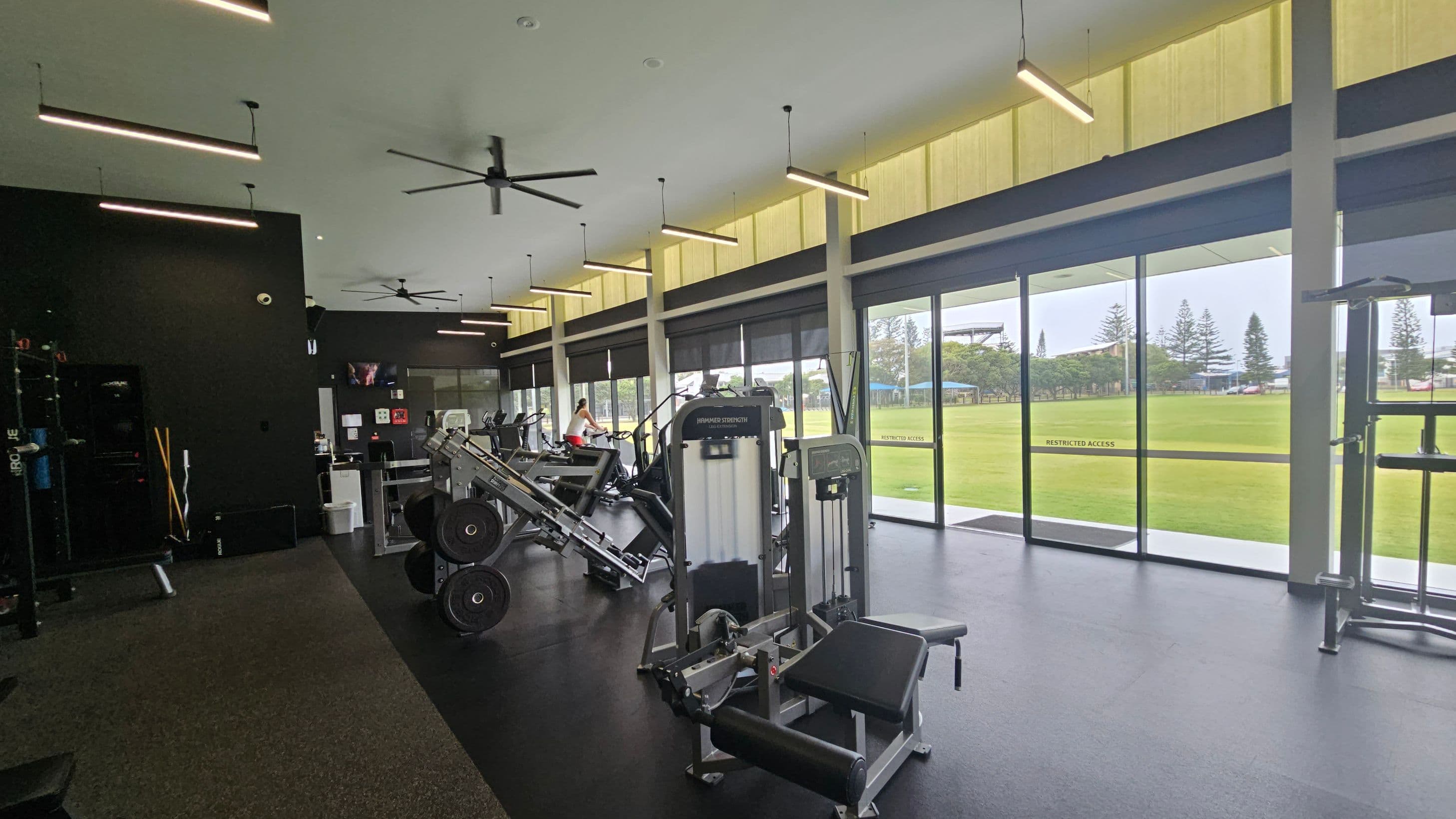 A modern gym interior with exercise equipment on the left, including weight machines and a leg press. The floor is dark with a green section. Large windows on the right provide a view of a grassy field and trees outside. The ceiling is high with linear lights and ceiling fans. A person is using a stationary bike in the background.