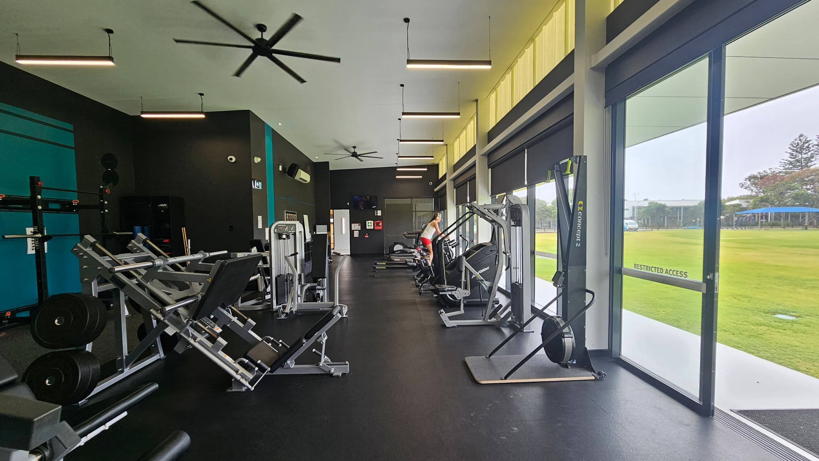 A gym interior with various exercise equipment. On the left, there are weight machines and racks. In the middle, a person is using a treadmill. On the right, large windows and glass doors provide a view of a grassy outdoor area. The floor is dark, and the ceiling has several hanging lights and ceiling fans.