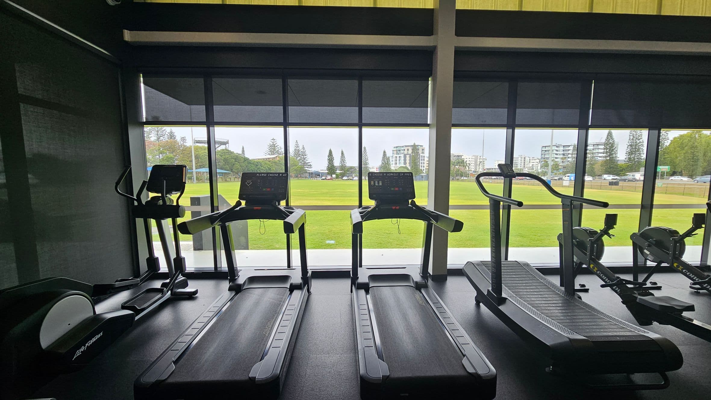 A gym interior with several treadmills and exercise machines lined up in front of large windows. The windows reveal a view of a green field and buildings in the background. The floor is dark and the ceiling is not visible. The equipment includes two standard treadmills, an elliptical machine on the left, and a curved treadmill on the right. The outdoor scene includes trees and a clear sky.