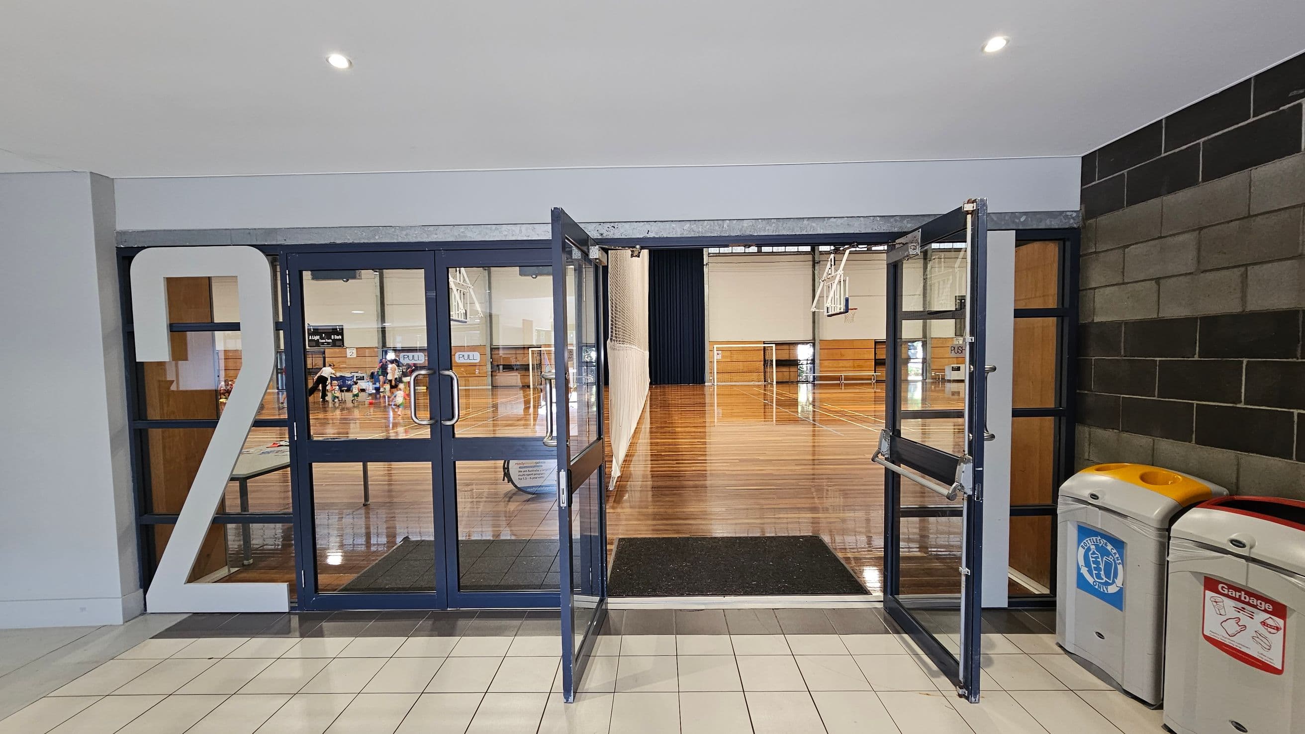 A hallway with a large number "2" on the left side of a glass door entrance. The doors are open, leading into a gymnasium with a polished wooden floor. In the background, there are basketball hoops and a net. On the right side, there are two bins: one for bottles and cans, and another for general garbage. The floor in the foreground is tiled, and the ceiling is plain with recessed lighting.