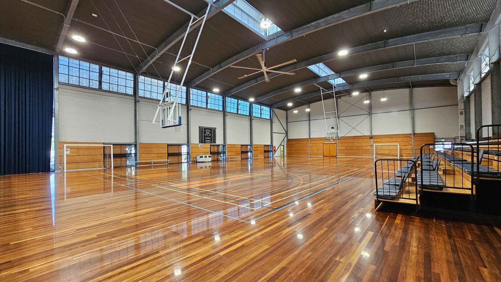 A spacious indoor sports hall with a polished wooden floor. On the left, there is a basketball hoop and a goalpost. The right side features tiered seating with black railings. The ceiling is high with large skylights and industrial lighting. The walls are lined with wooden panels and windows near the top. A scoreboard is visible in the centre background.