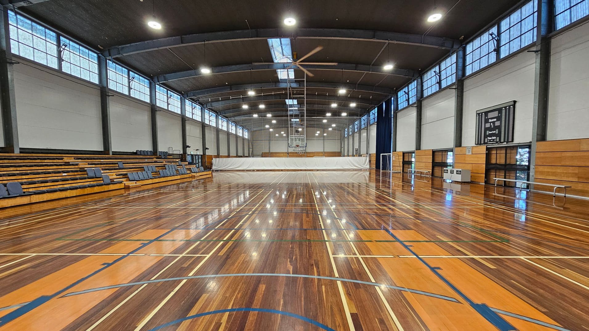A spacious indoor sports hall with a polished wooden floor marked for various sports. On the left, there are tiered wooden bleachers with blue seats. The right side features a scoreboard and some benches. The ceiling is high with large lights and a central fan. Windows line the upper walls, allowing natural light to enter. A white curtain divides the space at the far end, with a basketball hoop visible above it.