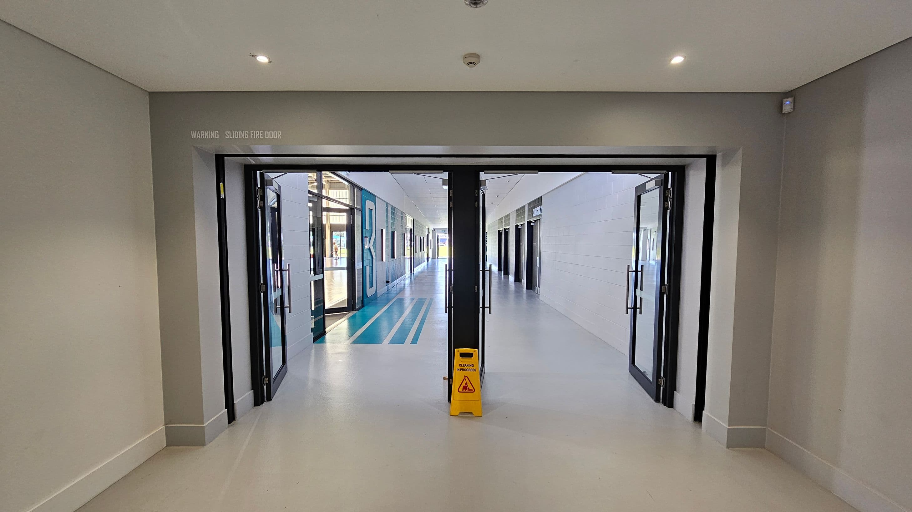 A corridor with a sliding fire door in the centre, partially open, leading to a hallway. The floor is light grey, and a yellow "Caution: Cleaning in Progress" sign is placed in front of the door. The walls are light-coloured, and the ceiling has recessed lighting. The hallway beyond has blue and white striped flooring, with glass doors and windows on the left and white panelled walls on the right.
