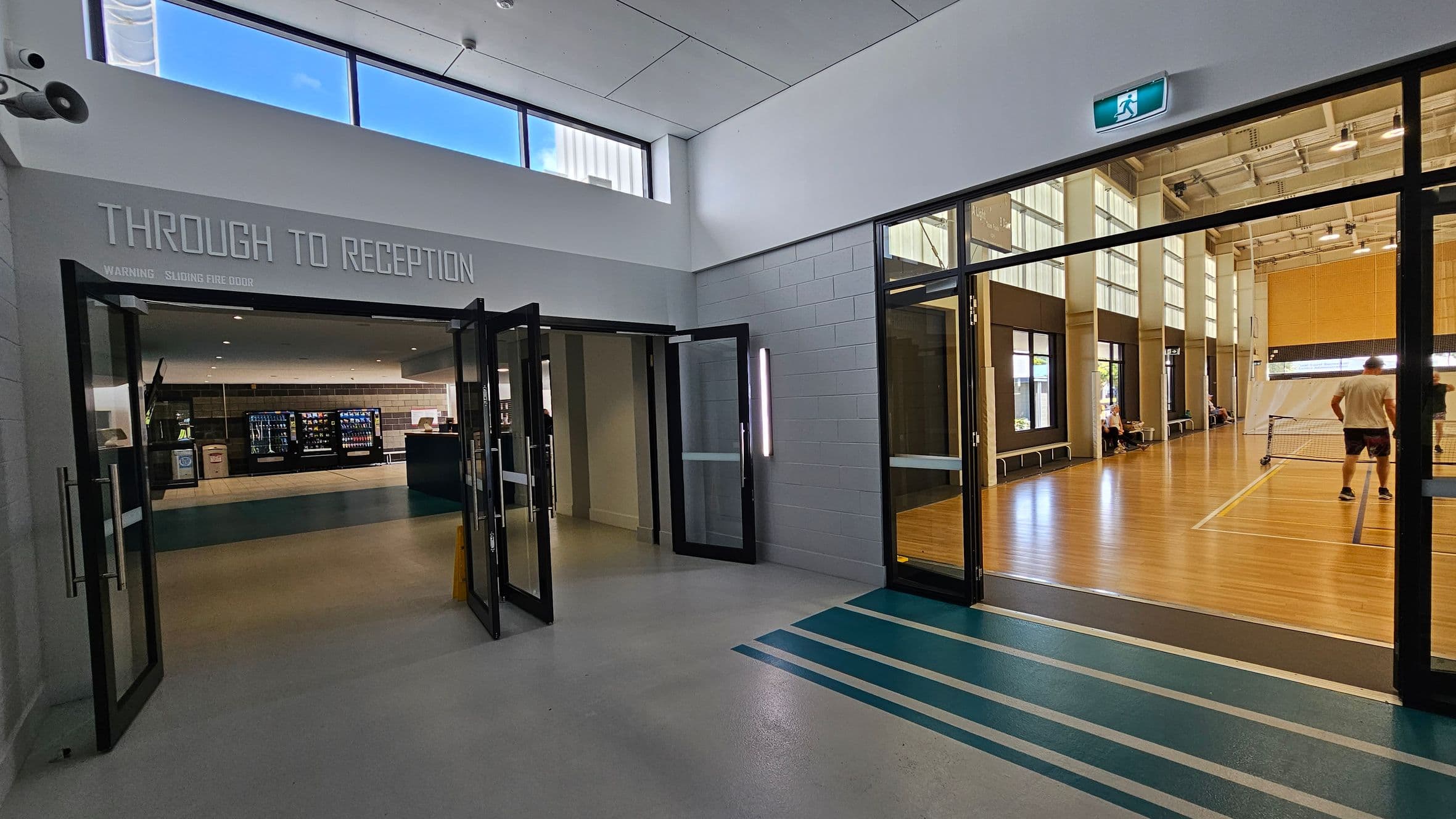 A hallway with a polished floor leads to two areas. On the left, glass doors open to a reception area with vending machines and a counter. On the right, another set of glass doors reveals a sports hall with wooden flooring, where people are playing a game. Above, a high ceiling with windows allows natural light in. An exit sign is visible above the doors to the sports hall.