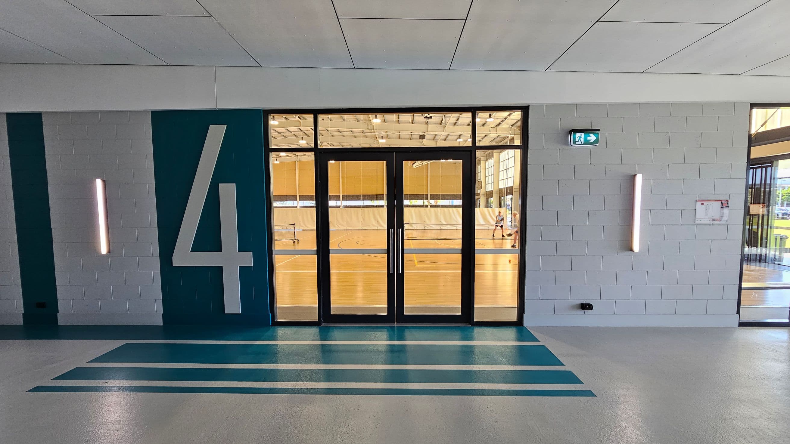 A hallway with a large number "4" on the left wall, painted in white on a teal background. The floor has teal stripes leading to a set of glass double doors. Through the doors, a gymnasium with a wooden floor is visible, where people are playing sports. The walls are light grey with vertical light fixtures on either side of the doors. An emergency exit sign is above the right light fixture. The ceiling is white with rectangular panels.