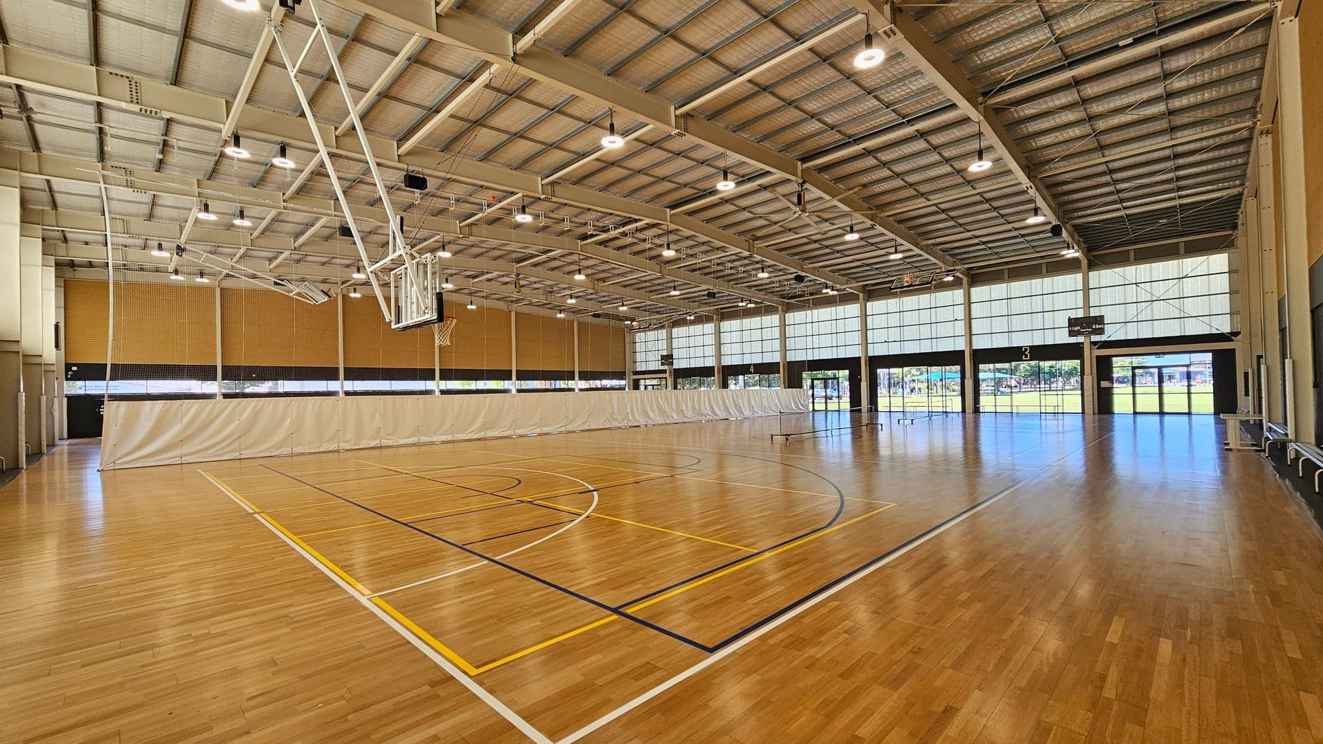 A spacious indoor sports hall with a polished wooden floor marked for basketball. On the left, a basketball hoop is suspended from the ceiling. The ceiling is high with exposed beams and numerous lights. A large white curtain divides the space in the middle. On the right, there are large windows and glass doors allowing natural light to enter, with a view of a grassy area outside. The walls are lined with light-coloured panels.