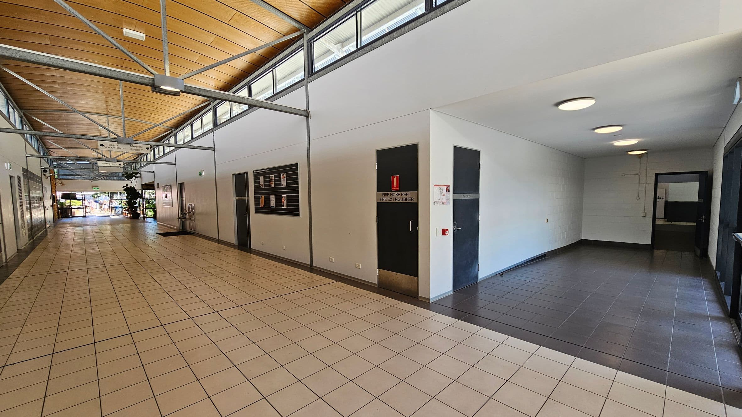 A hallway with a light brown tiled floor and a wooden panelled ceiling. On the left, there are doors and a notice board on the wall. The right side features a dark door labelled "Fire Hose Reel Fire Extinguisher" and another door marked "Plant Room". The ceiling has exposed metal beams and circular lights. At the end of the hallway, there is a glass door leading outside, with some greenery visible through it.