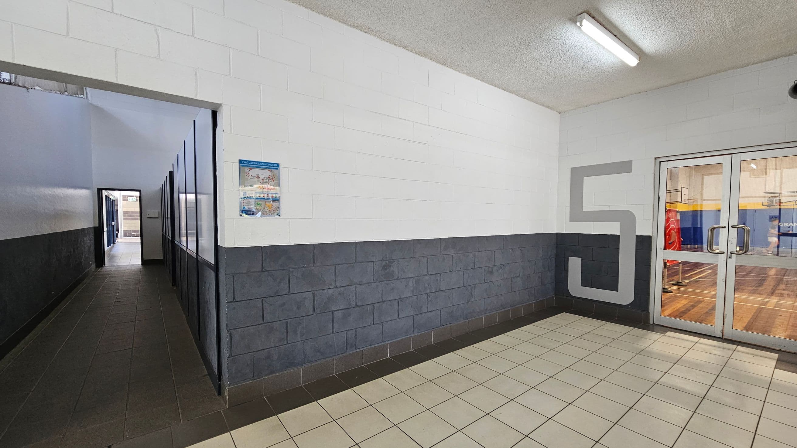A hallway with a tiled floor and a white ceiling. On the left, a narrow corridor with dark tiles and a series of windows or panels along the wall. On the right, a large number "5" is painted on the wall next to a set of glass double doors leading to a gymnasium with a wooden floor. A poster is mounted on the wall near the corridor entrance. The walls are painted white with a dark grey lower section. Fluorescent lights are mounted on the ceiling.