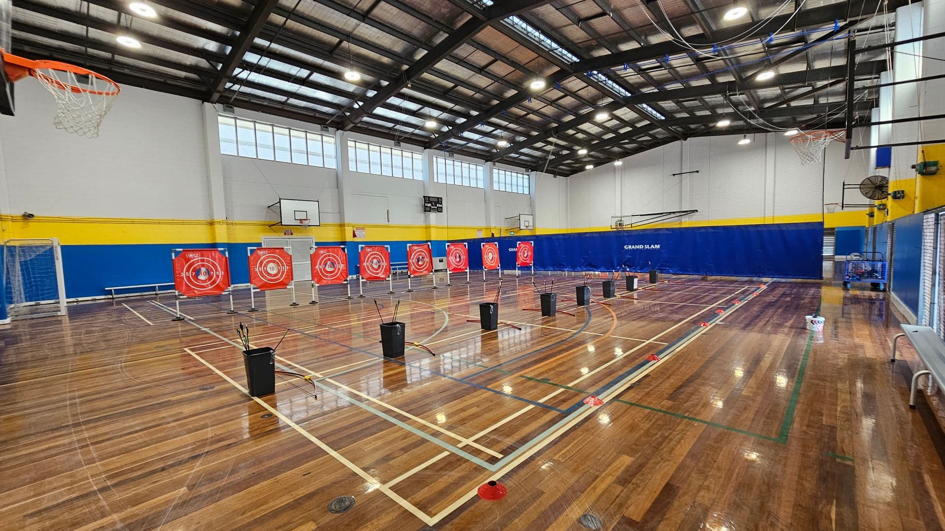 A gymnasium with a polished wooden floor and a high, metal-beamed ceiling. In the centre, there are several archery targets with red and white concentric circles. Black bins with arrows are placed in front of each target. On the left, a basketball hoop and a goalpost are visible. The walls are painted white with a blue and yellow stripe running horizontally. On the right, a bench and a blue wall with "GRAND SLAM" written on it can be seen.