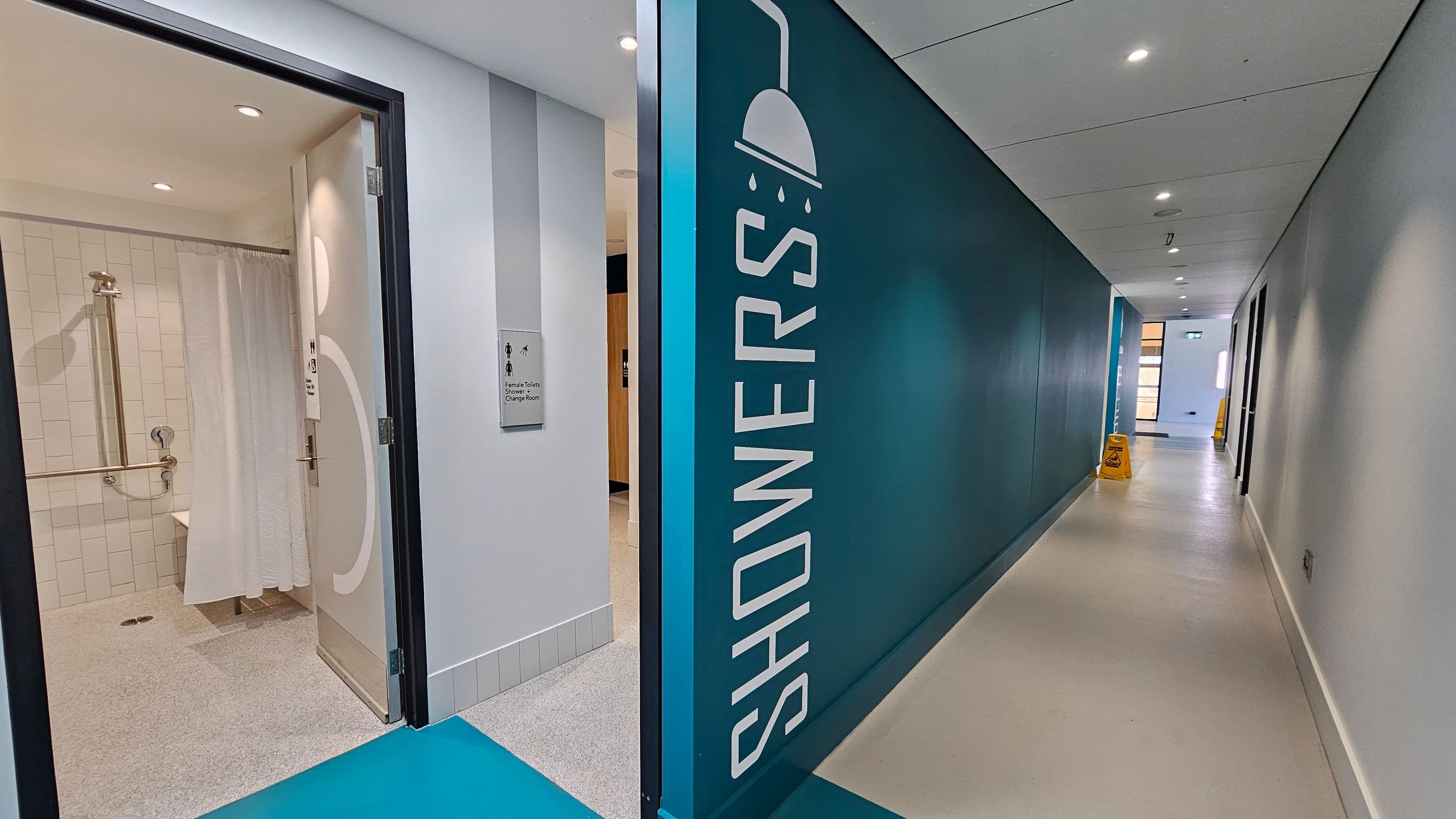 A corridor with a teal wall on the right featuring a large "SHOWERS" sign and a showerhead icon. On the left, an open door leads to a shower room with white tiles and a shower curtain. A sign next to the door reads "Female Toilets Change Room." The floor is light-coloured, and the ceiling has recessed lighting. Further down the corridor, a yellow caution sign is visible near a windowed area.