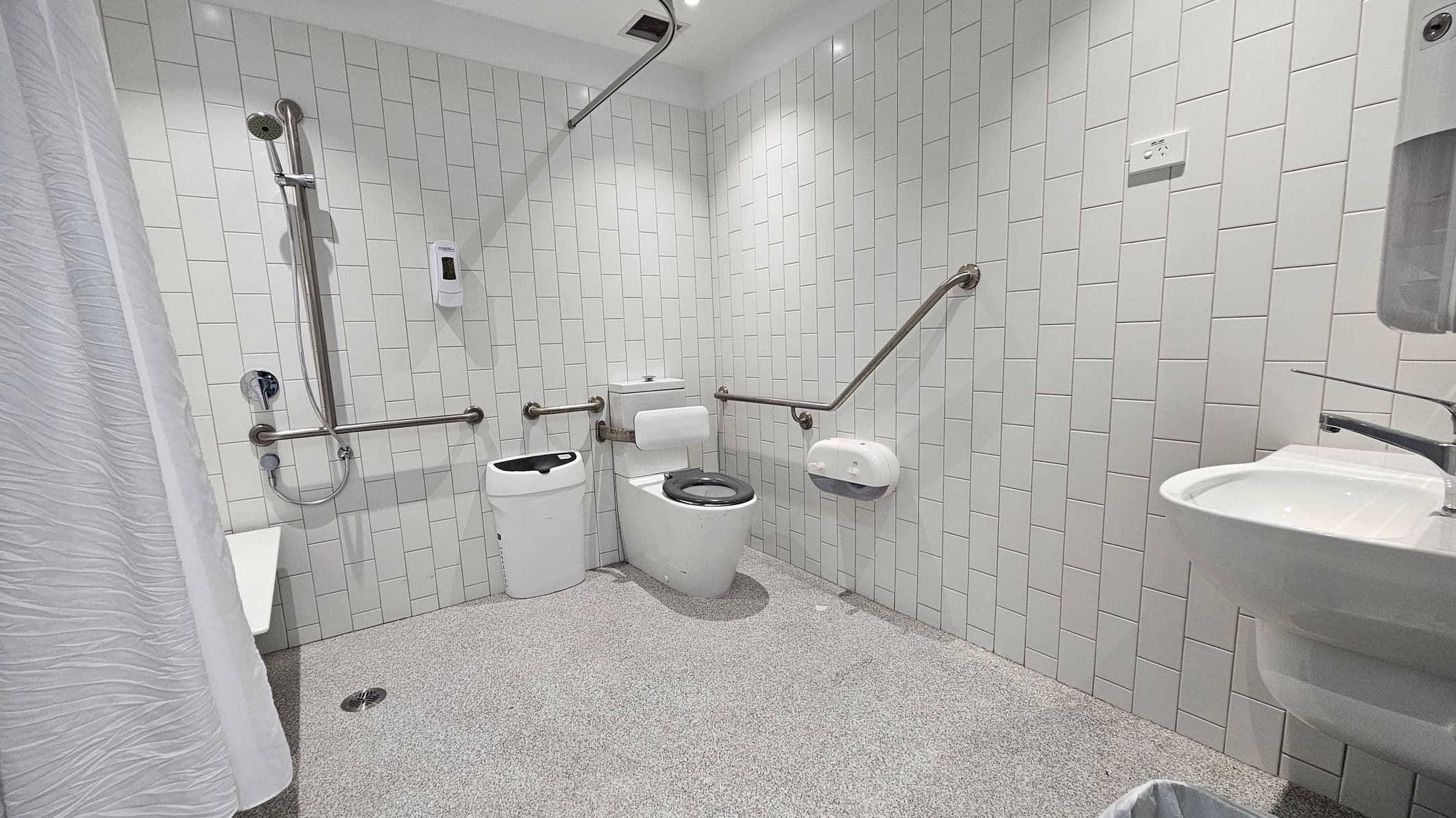 A modern, accessible bathroom with white tiled walls and a speckled grey floor. On the left, there's a shower area with a handheld showerhead and a white curtain. In the centre, a toilet with a black seat is flanked by grab bars. A sanitary bin is next to the toilet. On the right, a wall-mounted sink with a soap dispenser above it. The ceiling is white with recessed lighting.