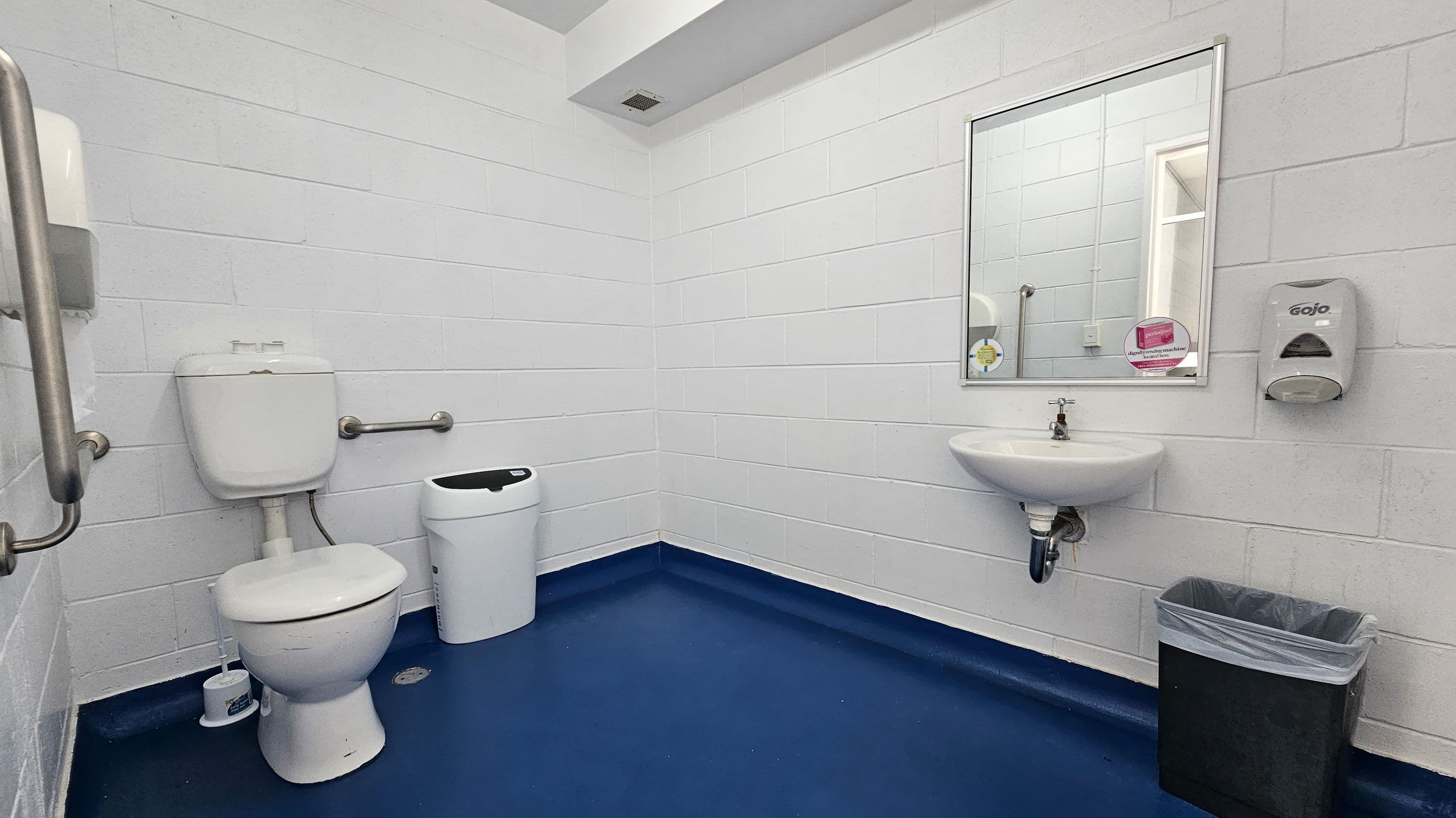 A public bathroom with white brick walls and a blue floor. On the left, there is a toilet with a grab bar and a small bin nearby. In the centre, a wall-mounted sink is below a rectangular mirror. To the right of the sink, a soap dispenser is mounted on the wall. Below the dispenser, there is a black waste bin with a liner. The ceiling is white with a small vent.