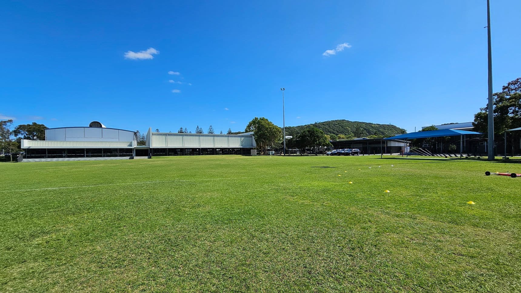 A large grassy field with a few scattered yellow cones in the foreground. In the background, there are several modern buildings with white facades. To the right, a covered area with a blue roof is visible, along with a row of parked cars. A tall light pole stands near the centre, and trees are visible behind the buildings, with hills in the distance. The sky is clear with a few small clouds.