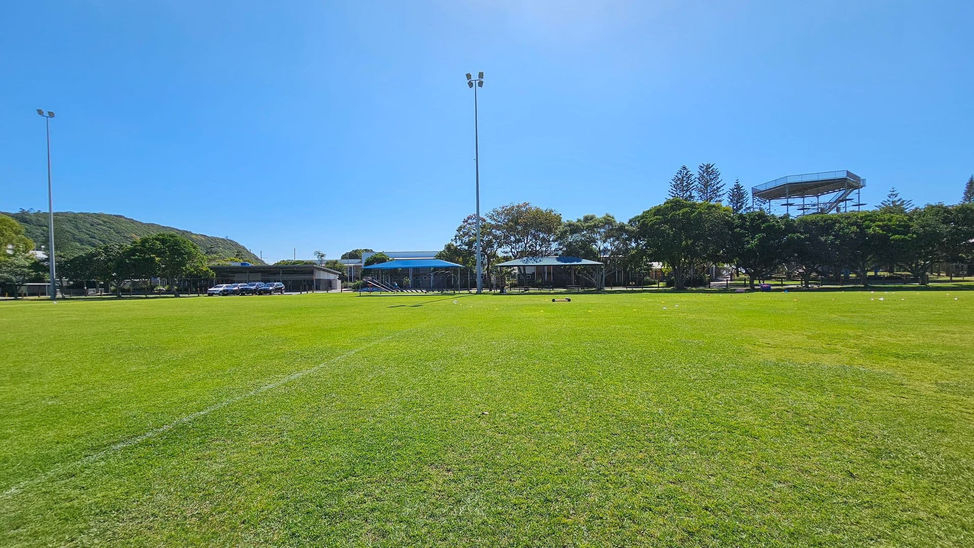 A large grassy field with a few scattered white lines. In the background, there are several trees and a row of buildings, including a structure with a blue roof. A tall light pole stands in the centre. To the right, there is a metal observation tower. A hill covered with greenery is visible on the left side. Several cars are parked near the buildings.