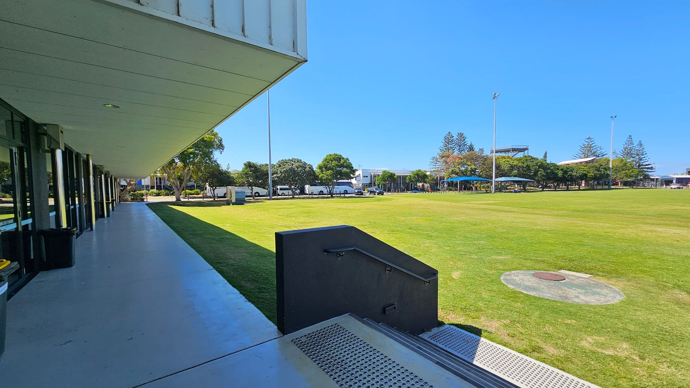 A wide, open grassy field is on the right, bordered by trees and several tall light poles. In the background, there are buildings and a row of parked vehicles. On the left, a modern building with large glass windows and a covered walkway extends along the edge of the field. The foreground features a set of steps with a metal railing leading down to the grass.