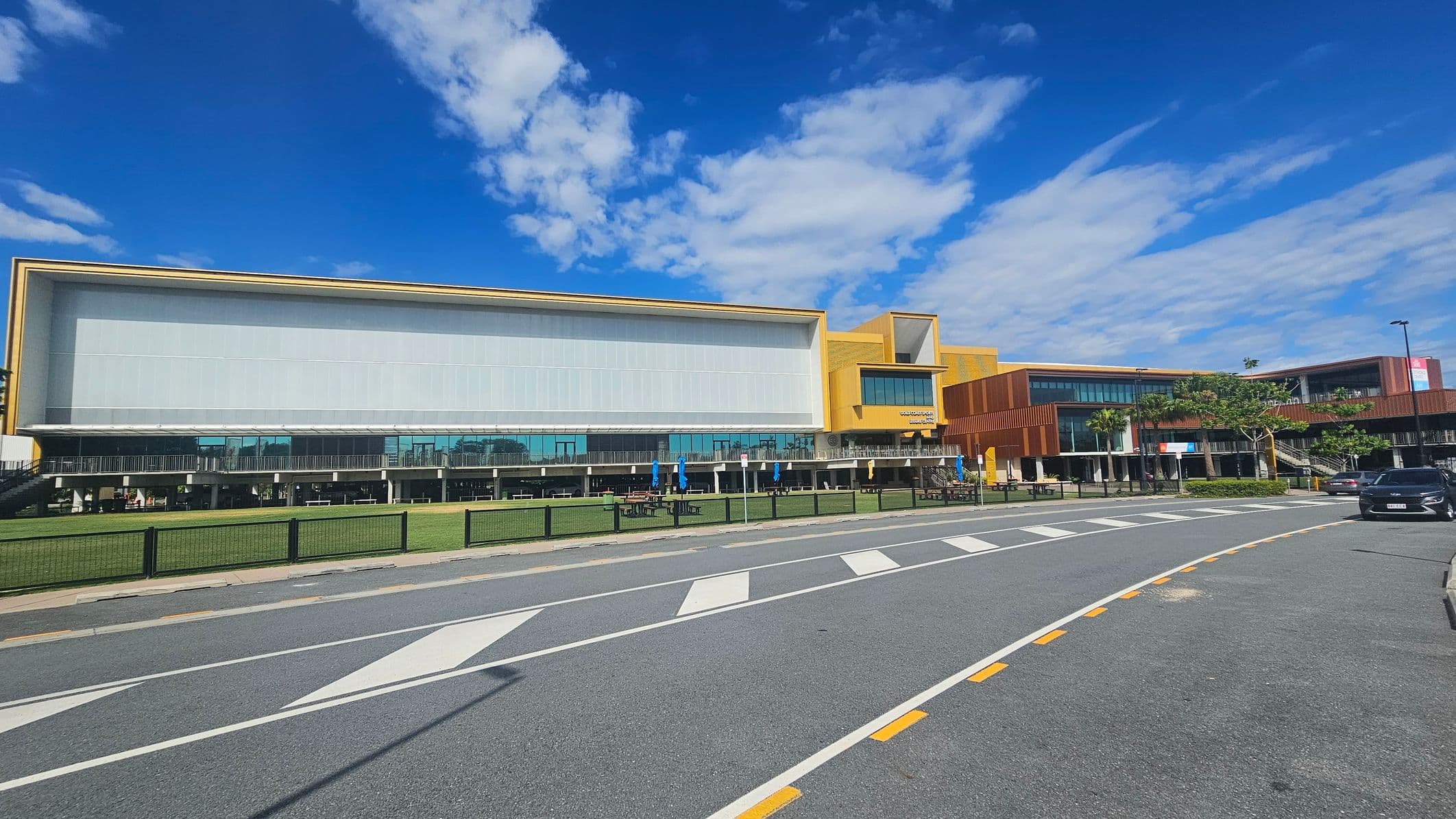 A modern building with a large glass facade and yellow accents on the right. The structure is two-storeyed with a mix of glass and wooden panels. In front, there's a grassy area with picnic tables and umbrellas. A road with marked pedestrian crossings runs parallel to the building, and a black car is parked on the right side. The sky is clear with some clouds.