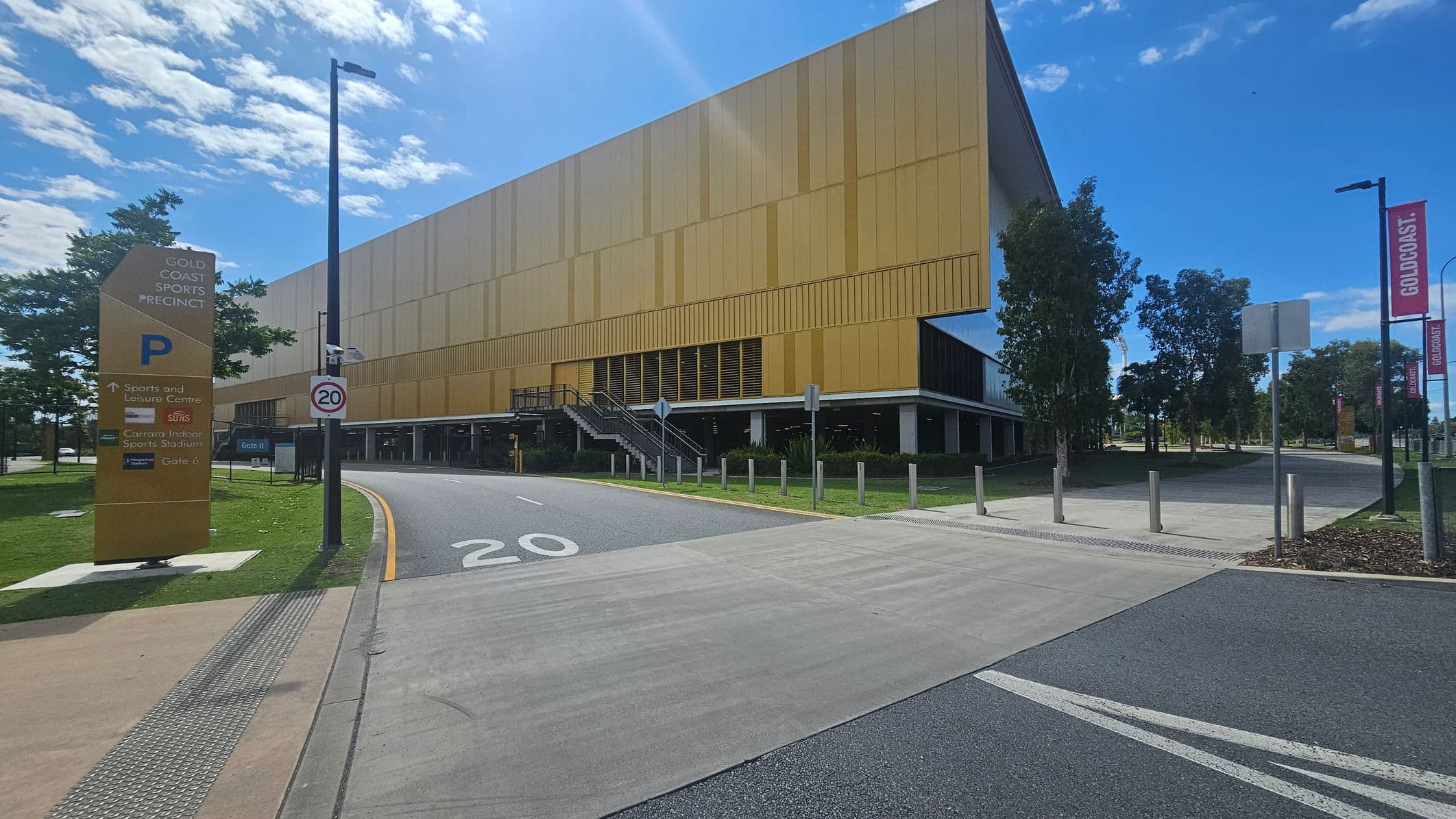 A large yellow building with a modern design is on the right, featuring a staircase leading to an entrance. In the foreground, a road with a speed limit of 20 is marked. On the left, a tall sign reads "Gold Coast Sports Precinct" with directions to various facilities. Trees line the area, and banners with "Gold Coast" are visible on lamp posts. The sky is clear with some clouds.