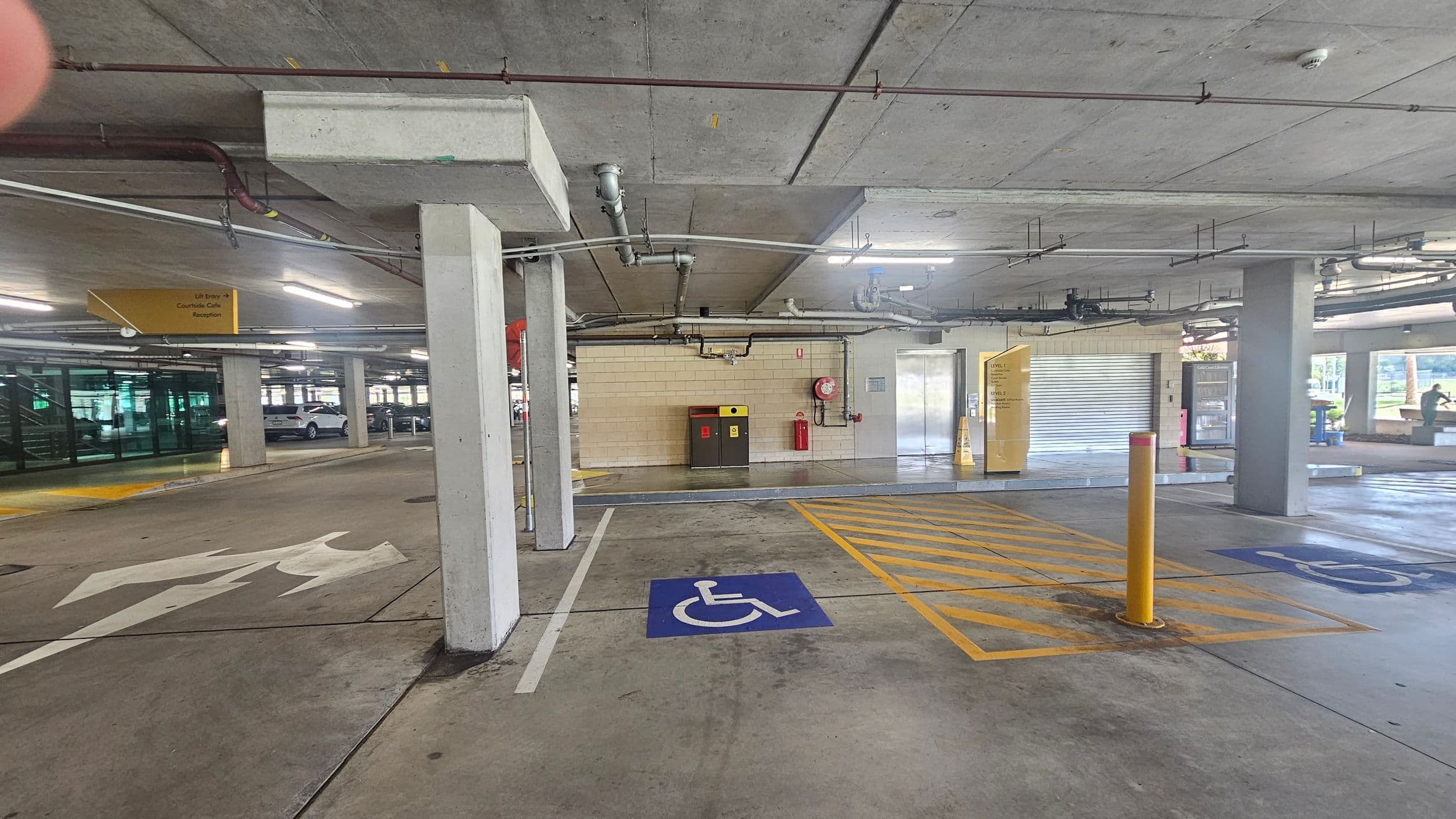 Underground car park with concrete ceiling and floor. On the left, there are parking spaces and a sign indicating directions. In the centre, a blue disabled parking space with yellow crosshatch markings and a yellow bollard. On the right, a wall with a fire extinguisher, a closed roller door, and a glass entrance. Pipes and lights are visible on the ceiling.