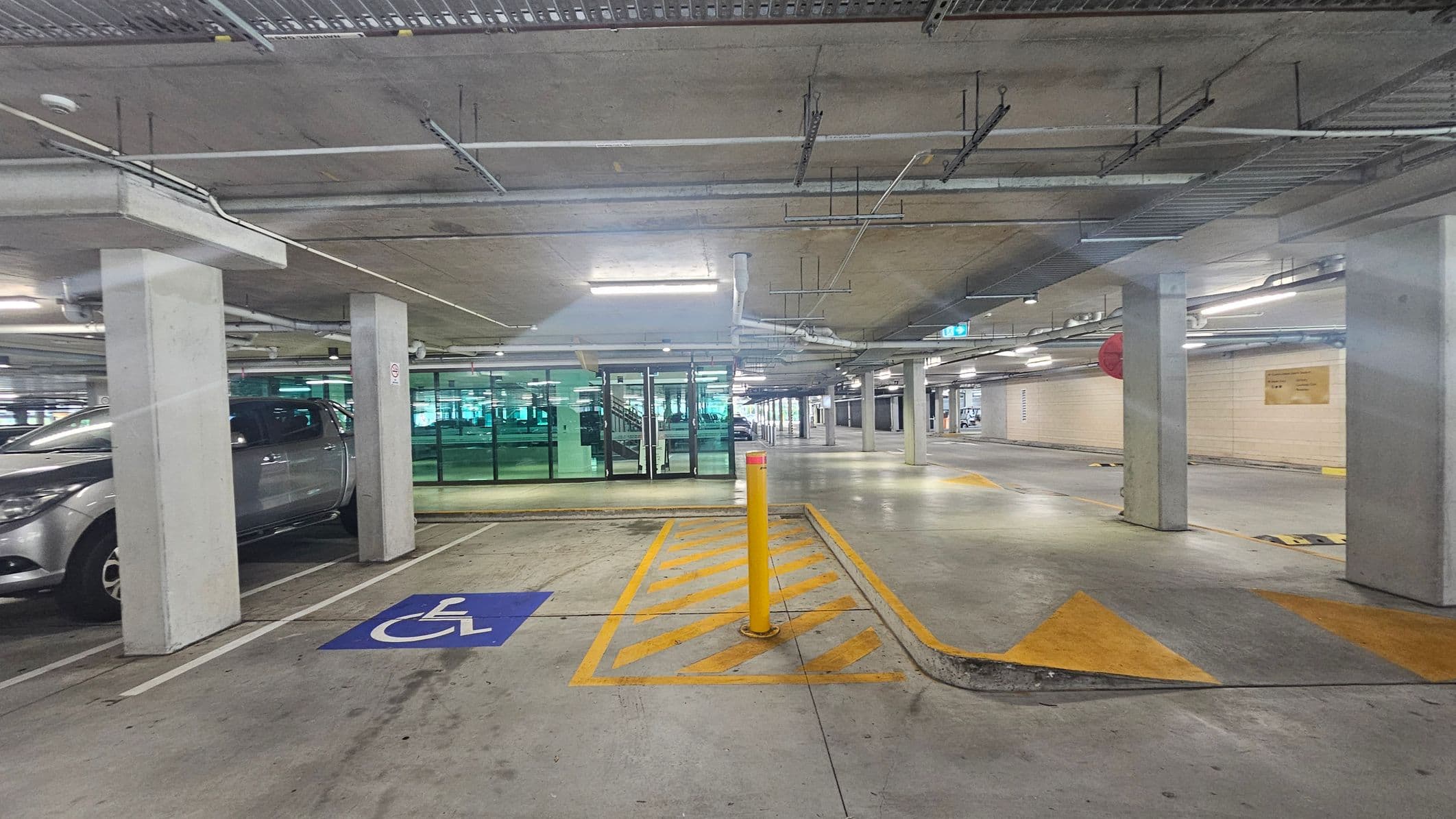 Underground car park with concrete pillars and a low ceiling. On the left, a silver vehicle is parked next to a blue disabled parking sign on the ground. In the centre, a yellow bollard stands in a hatched area. Glass doors and windows are visible in the background, leading to a stairwell. The floor is concrete, and the ceiling has exposed pipes and lighting fixtures. The right side shows an open space with directional arrows on the floor.