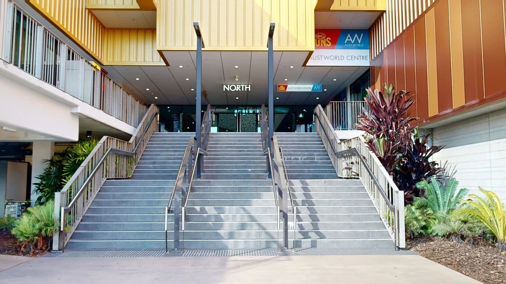 Wide staircase leading up to a building entrance with a sign reading "NORTH". The stairs are flanked by metal railings. On the left, there are green plants and a white wall. On the right, there are more plants, including tall red and green foliage, next to a brown panelled wall. Above the entrance, there are signs for various businesses. The ceiling above the stairs is flat with recessed lighting.