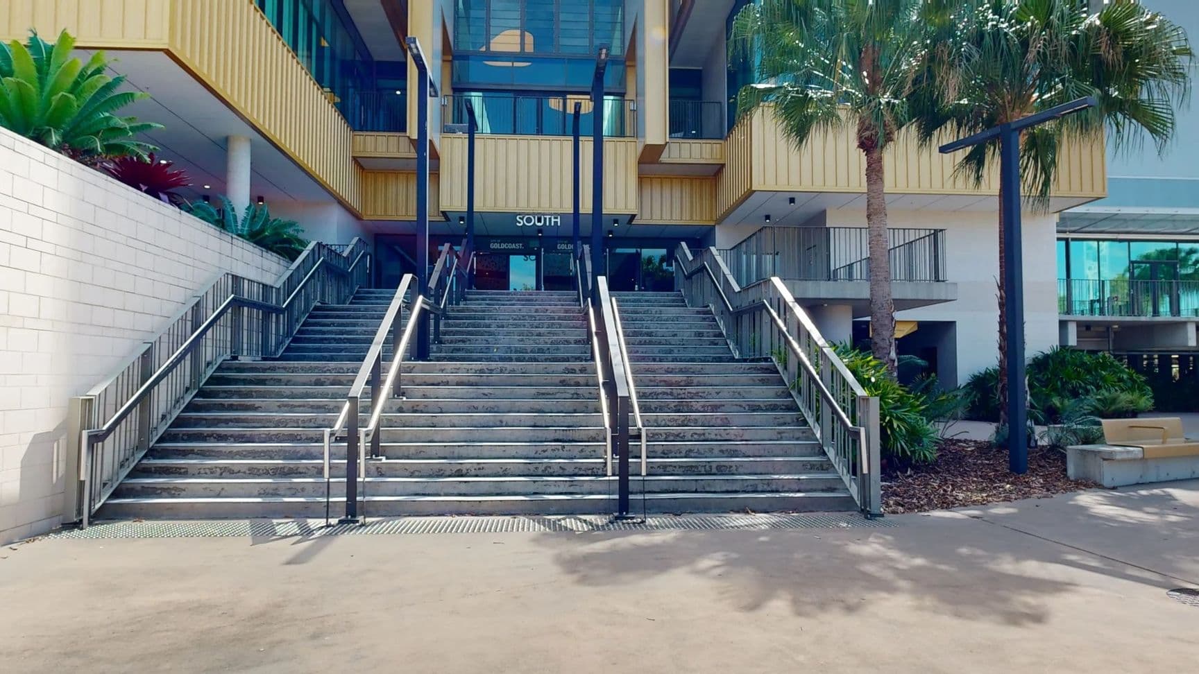 Wide concrete steps lead up to a building entrance marked "SOUTH". The steps are flanked by metal railings and bordered by lush green plants on both sides. On the right, a tall palm tree stands next to a modern streetlight. The building features a yellow facade with large glass windows. The ground is paved, and there are shadows cast by the trees and structures.