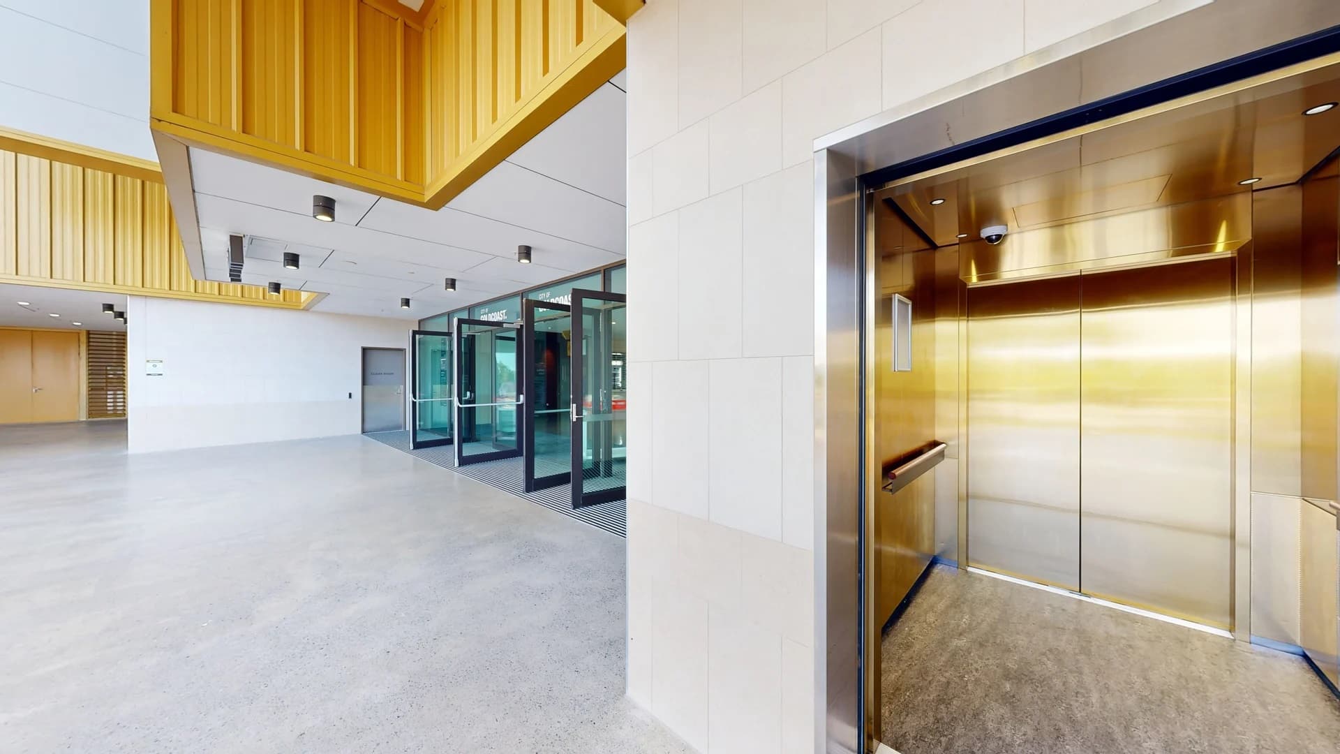 On the right, there's an open lift with shiny metallic walls and a grey floor. In the middle, a spacious lobby area with a light grey polished floor. On the left, a series of glass doors with metal frames lead to another area. The ceiling features white panels with black recessed lights, and there are wooden accents along the upper walls.